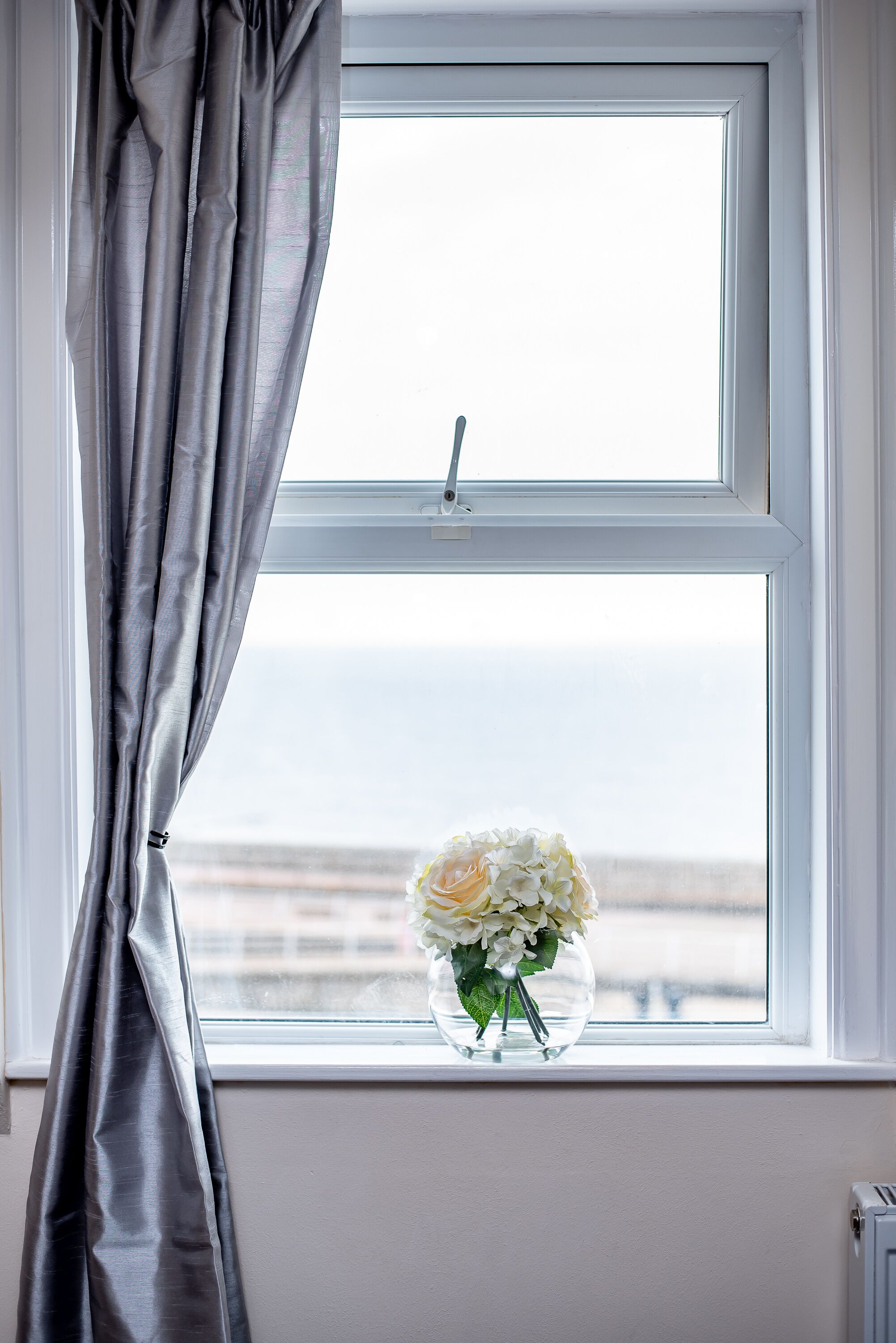A window with a view of the sea, a vase with flowers, and grey curtains.