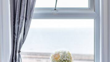 A window with a view of the sea, a vase with flowers, and grey curtains.