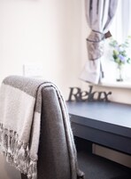 A chair with a grey blanket and tassels, a dark desk with a 'Relax' sign, and a window with curtains and a plant.
