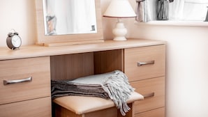 A wooden vanity with a mirror, a lamp, and a small bedside table with a blanket.