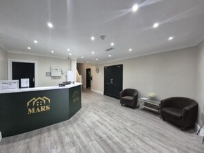 Zona de recepción con un mostrador identificado como 'MARS', un área de espera con dos sillones y una puerta que da a otra sala.