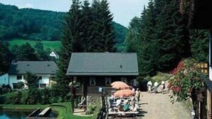 A wooden building with a sloped roof, a small pond, and a wooden deck with umbrellas. - Wolffhotel (Kopp)