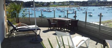 En terrasse med udsigt over en havn fyldt med både, et bord, stole og en stor parasol der giver skygge.