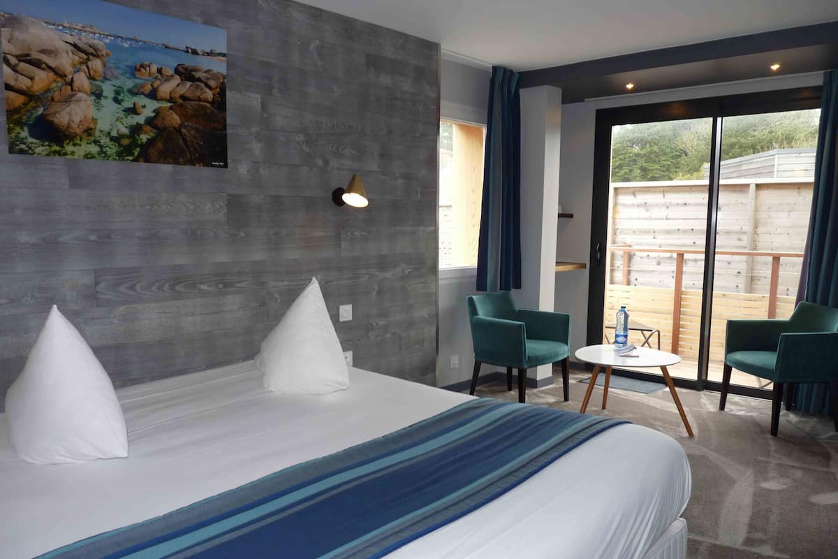 Habitació d'hotel moderna amb un llit gran, dues cadires, una taula petita i un balcó amb vistes a l'exterior.