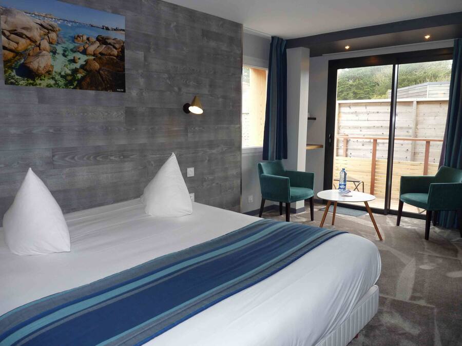 A modern hotel room with a large bed, two chairs, a small table, and a balcony with a view of the outdoors.
