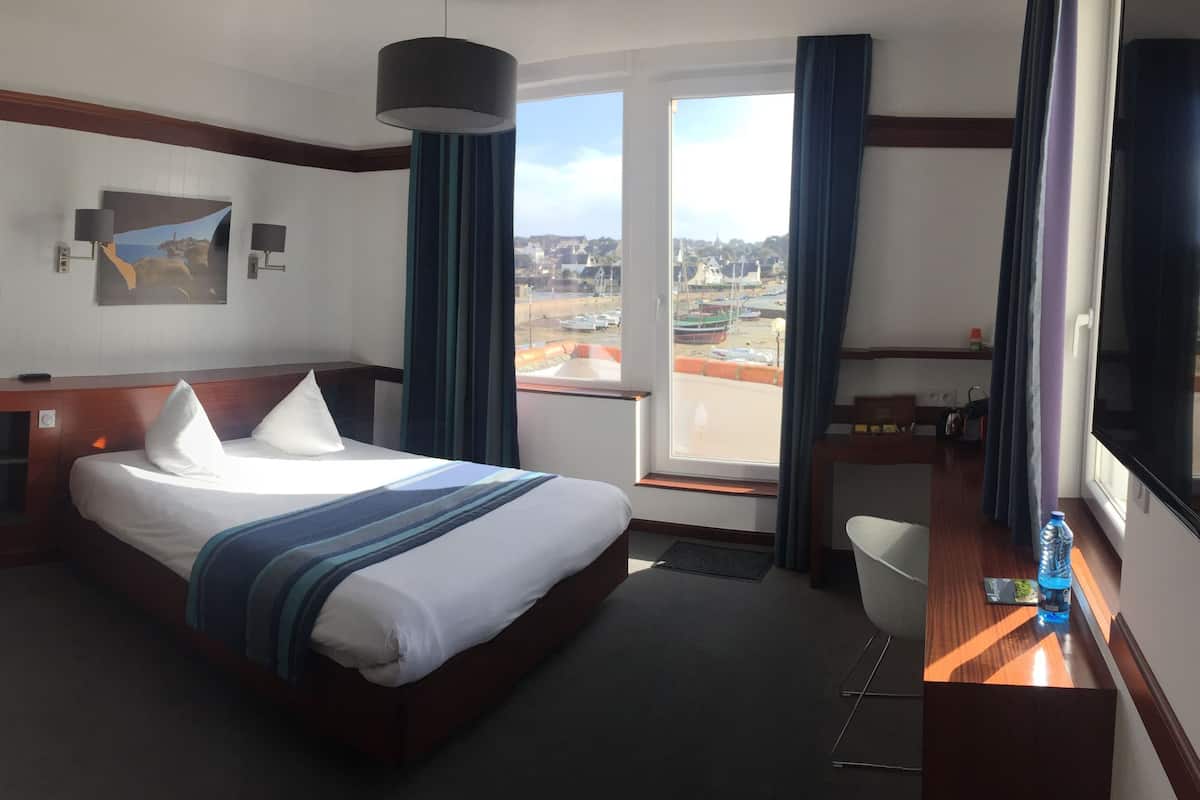 Habitació d'hotel amb un llit, un sofà, una taula de treball i vista a la ciutat a través de grans finestres.