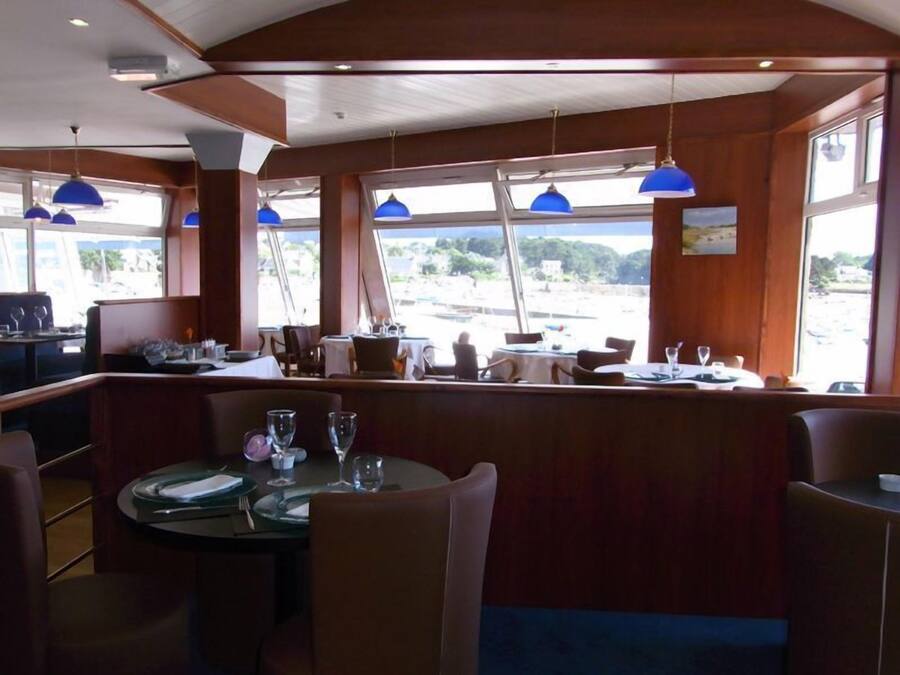 A restaurant with a view of the beach, featuring round tables, blue pendant lights, and wooden paneling.