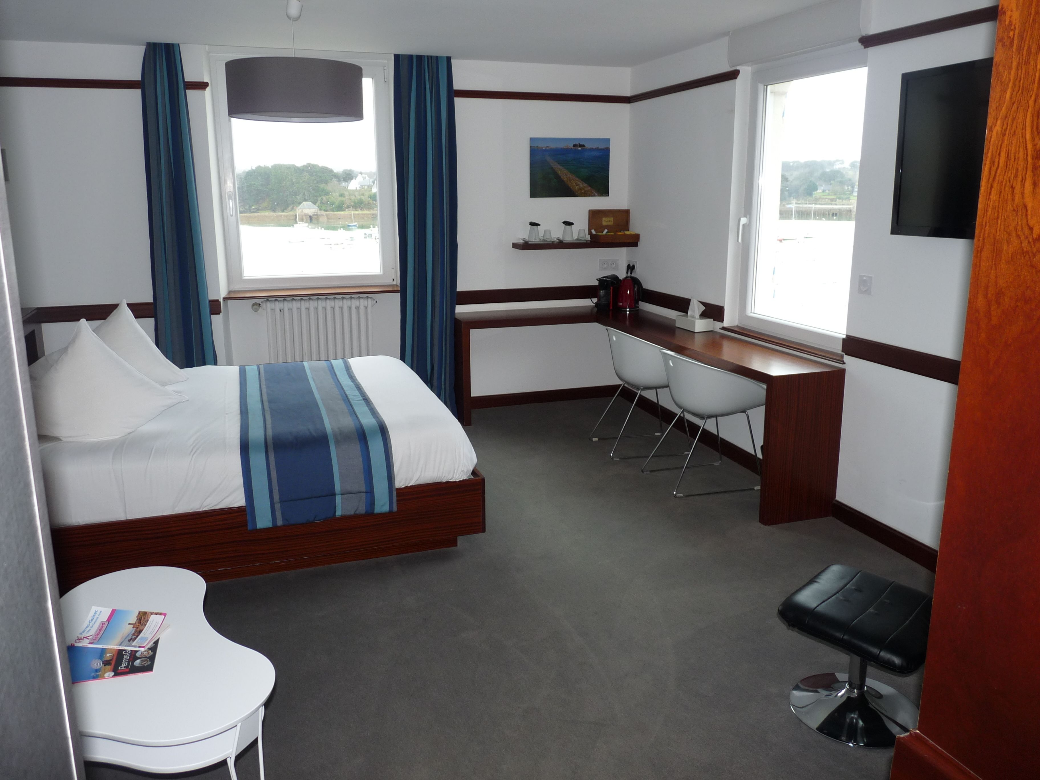 Ein Hotelzimmer mit einem Bett, einem Schreibtisch, zwei Stühlen, einem kleinen Tisch, einem Fernseher und einem Fenster mit Meerblick.