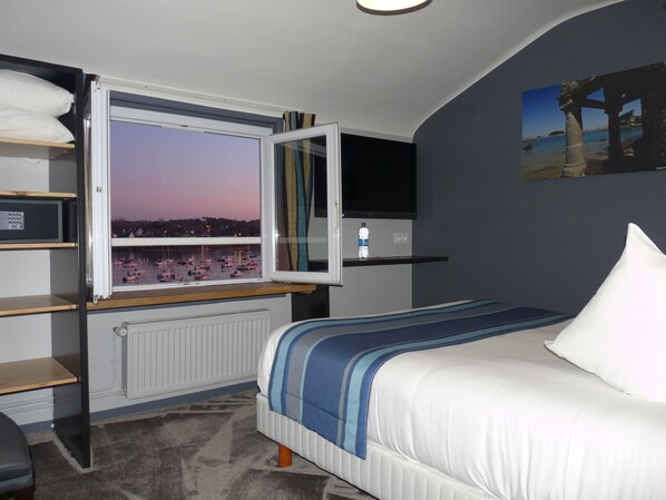 A bedroom with a bed, a window with a view of boats, a shelf with a bottle, and a TV on the wall.