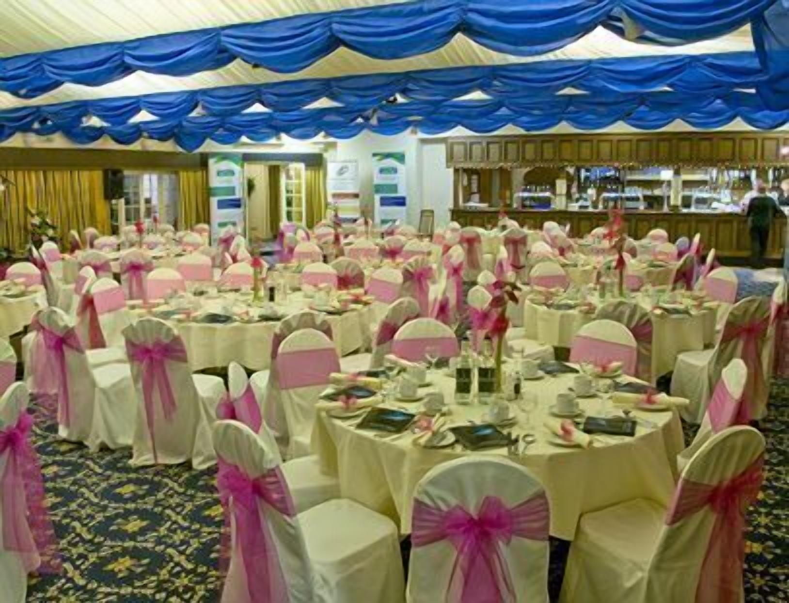 Un salón de banquetes con mesas redondas cubiertas con manteles blancos y cintas rosas, dispuestas en filas.
