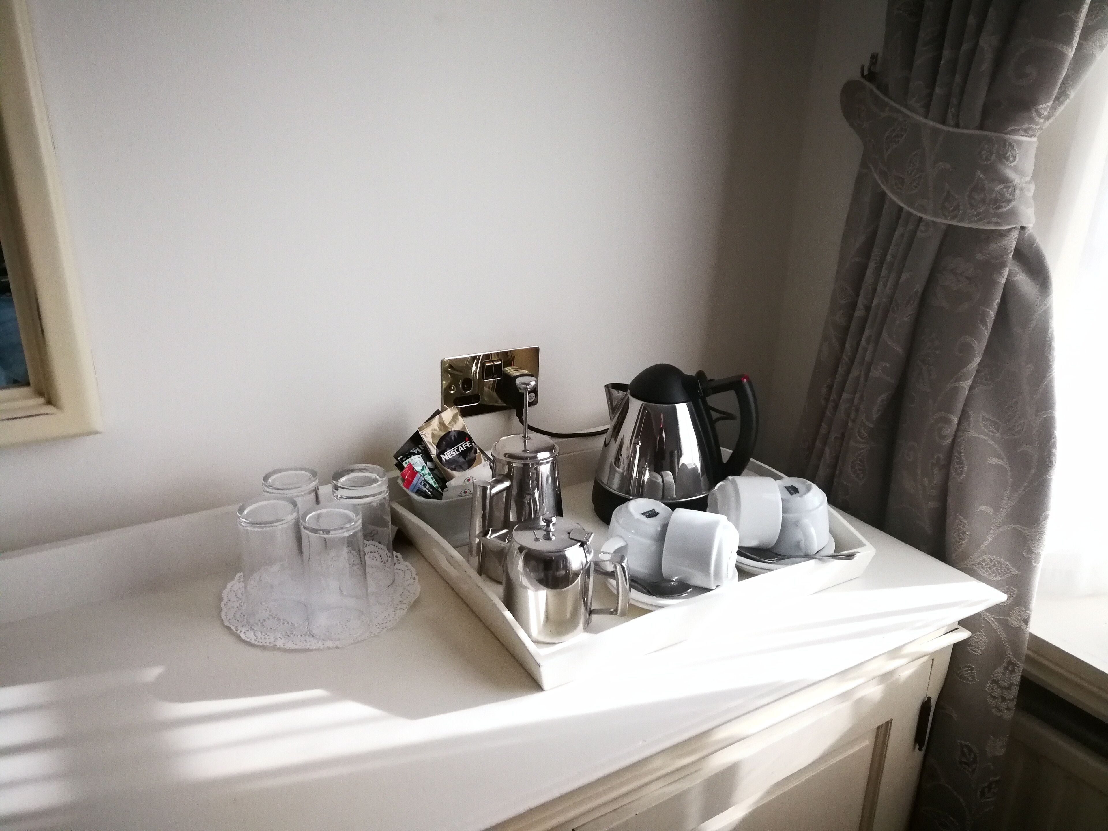 Bandeja de desayuno con tazas de café, una cafetera y un paquete de Nescafé sobre una mesa blanca cerca de una ventana con cortinas.