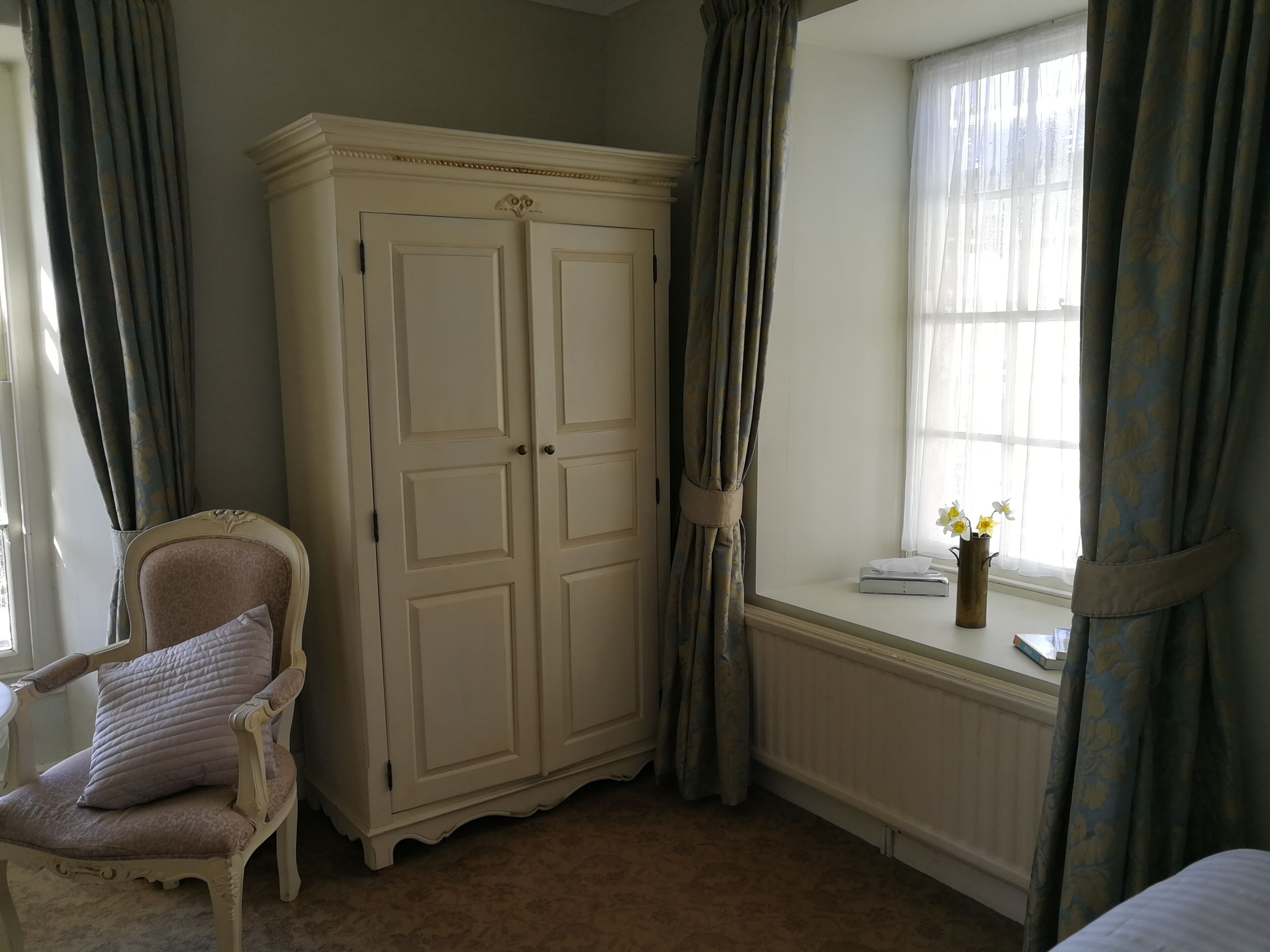 A room with a large white wardrobe, a cushioned chair, a window with curtains, and a radiator.