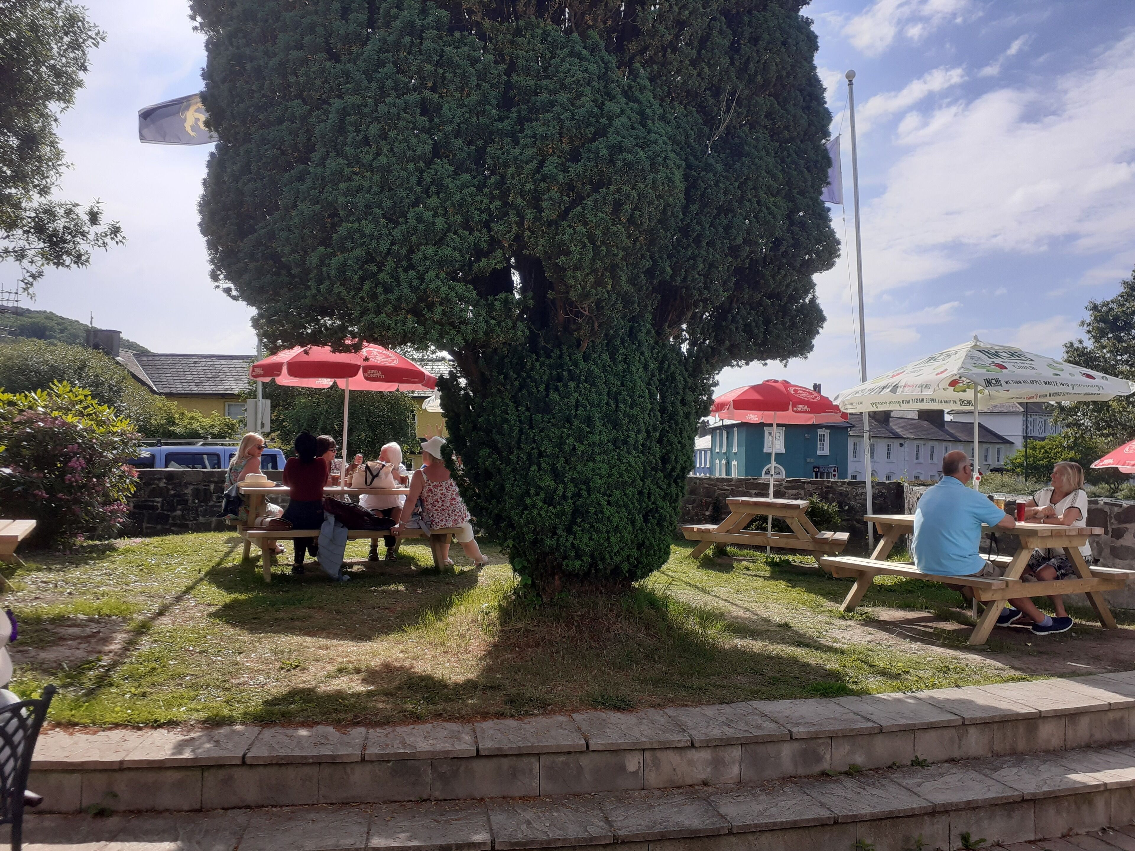 An outdoor seating area with picnic tables and red umbrellas, surrounded by greenery and a large tree.