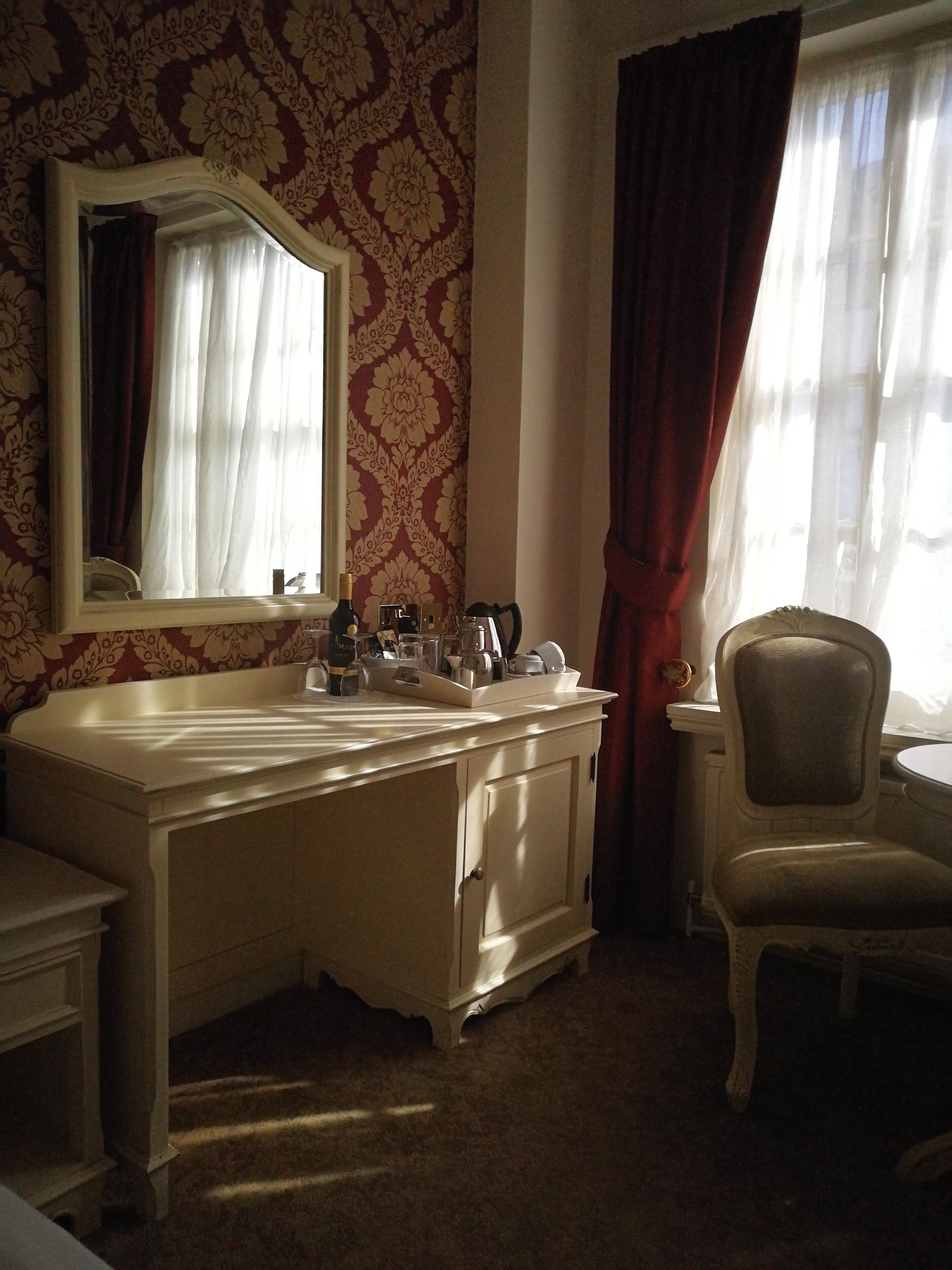 Une chambre d’hôtel avec une grande coiffeuse blanche, un miroir, une chaise et une fenêtre avec des rideaux.