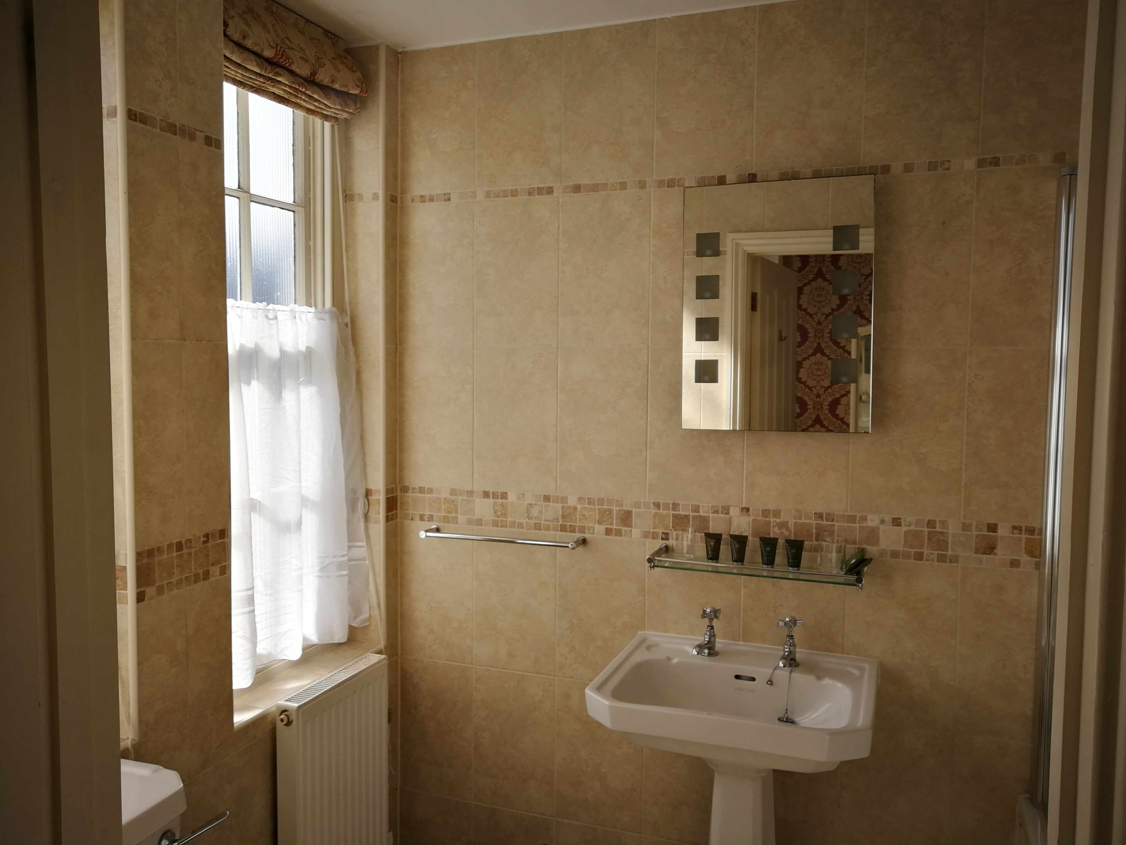 A bathroom with beige tiles, a window with a curtain, a sink, and a mirror.