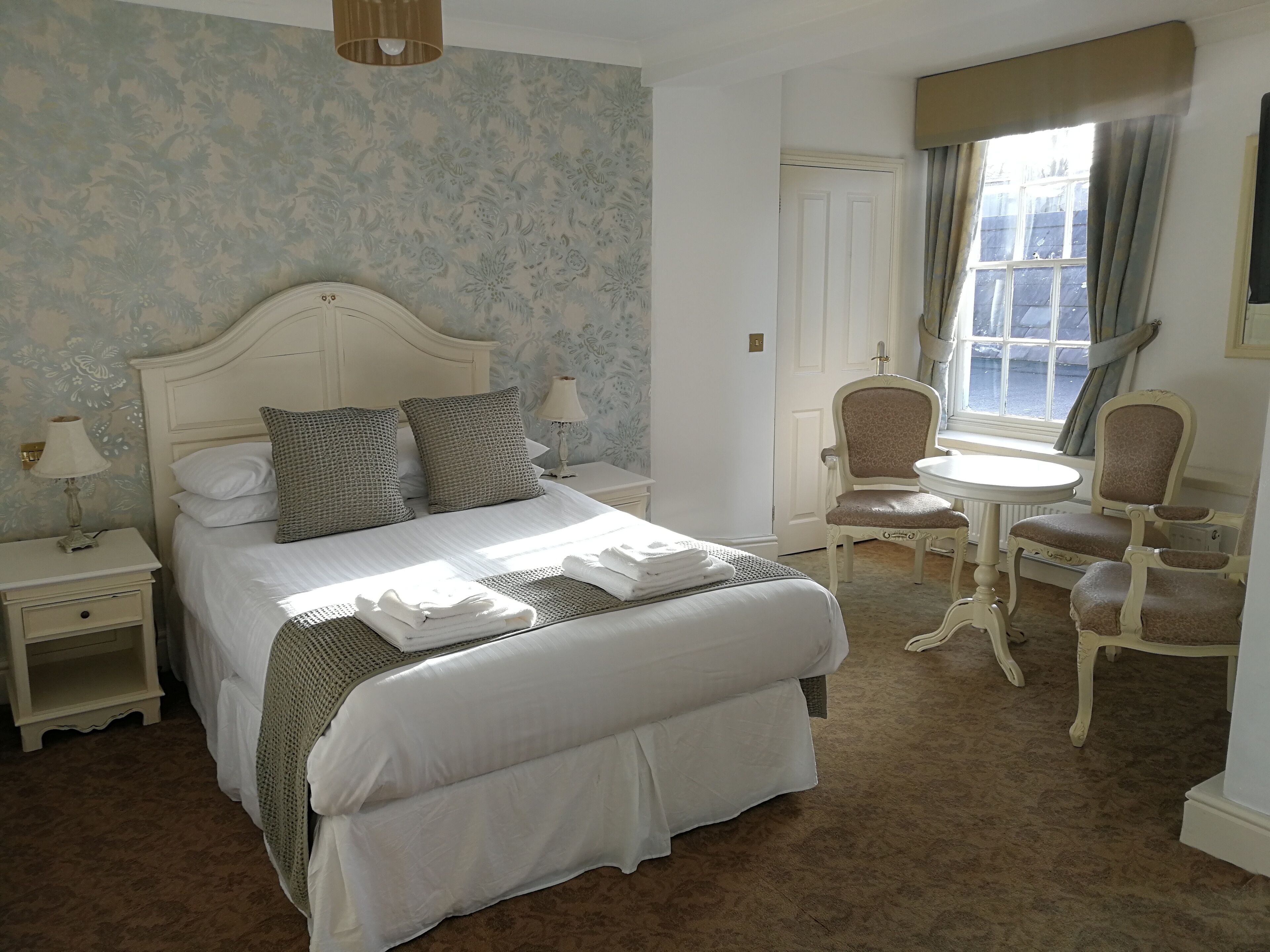 Une chambre d’hôtel bien aménagée, avec un grand lit, une petite table, deux chaises et une fenêtre avec des rideaux.