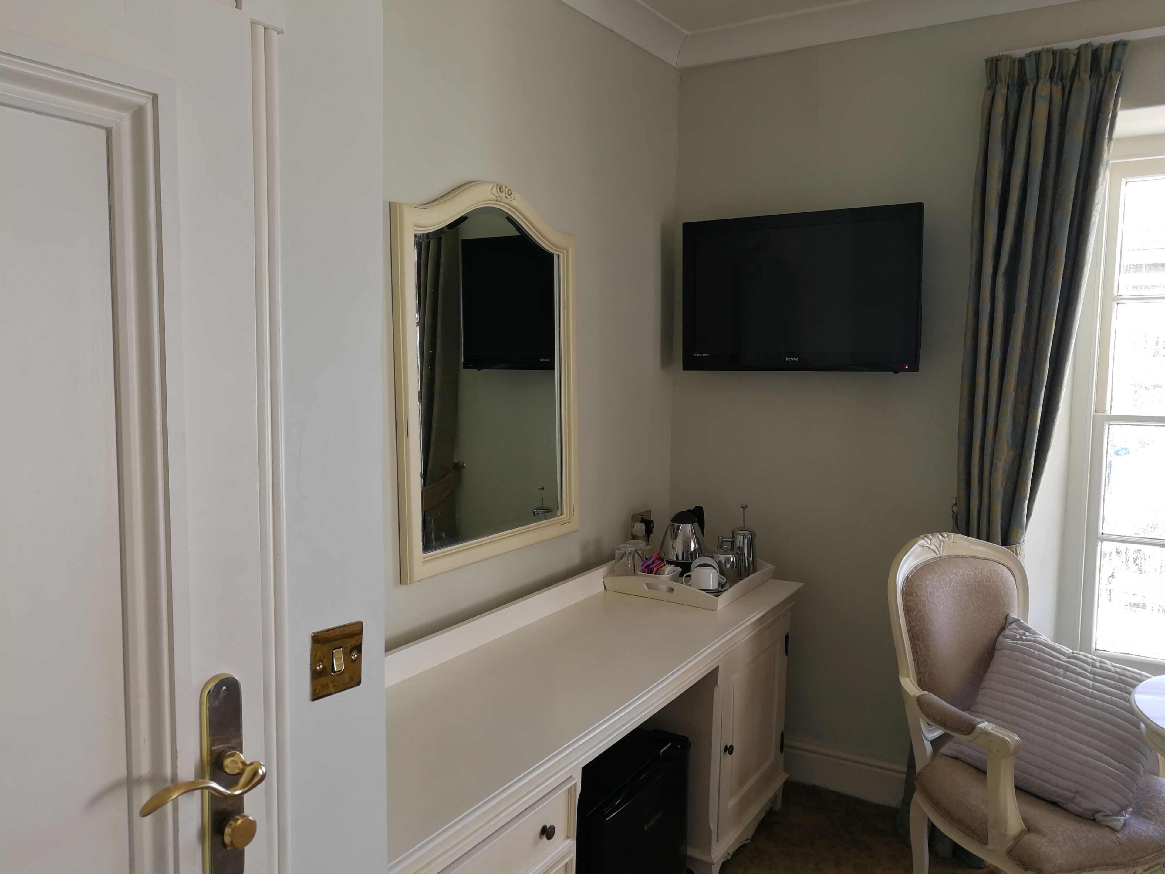 A room with a white door, a vanity with a mirror, a flat-screen TV, a chair, and a window with curtains.