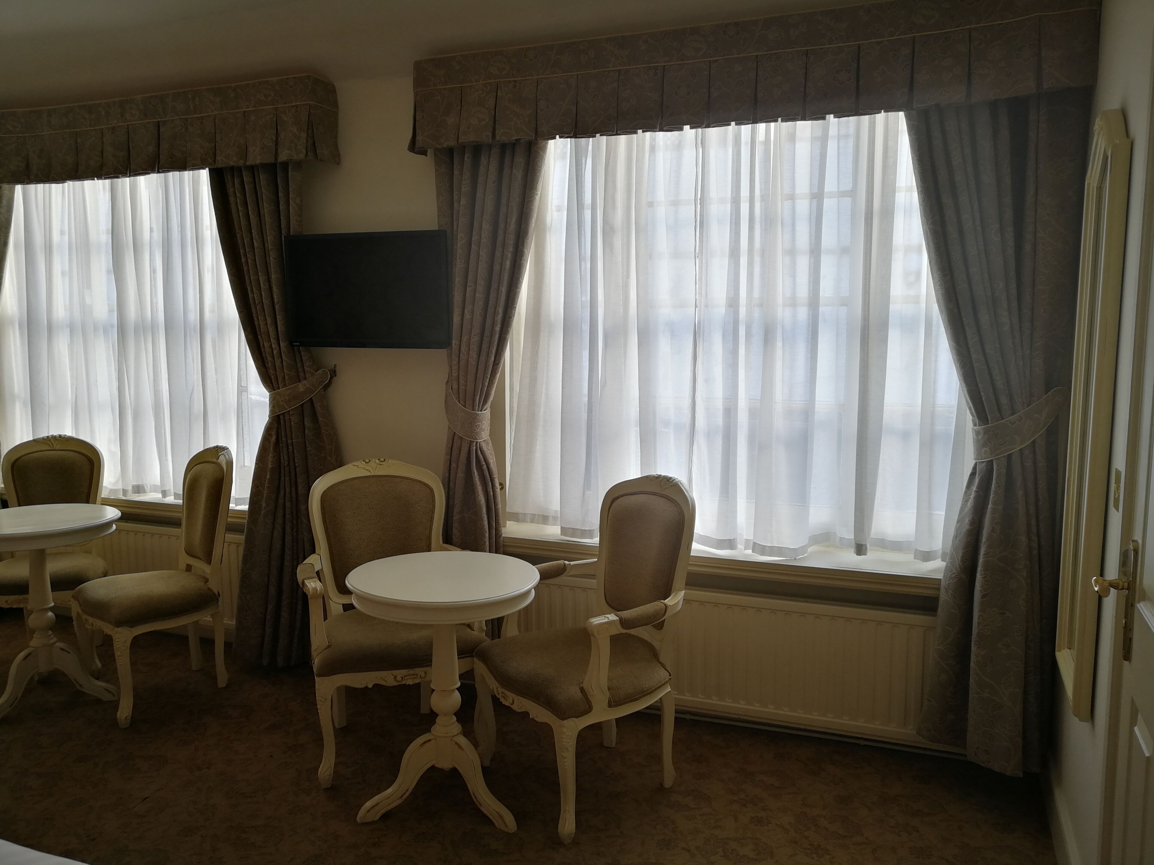 Une pièce avec deux tables rondes et quatre chaises, de grandes fenêtres avec des rideaux et un radiateur.