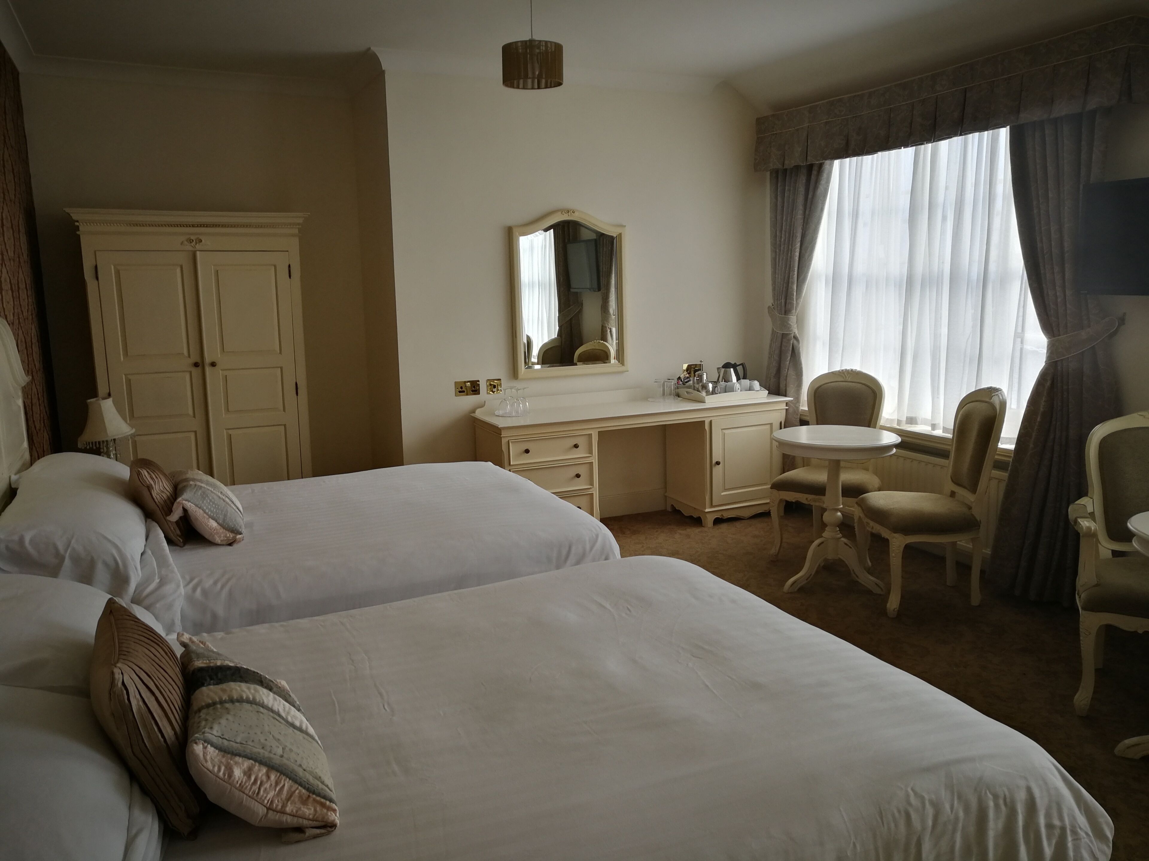 Ein Hotelzimmer mit zwei Betten, einem Schreibtisch, einem Stuhl, einem Spiegel und einem Fenster mit Vorhängen.