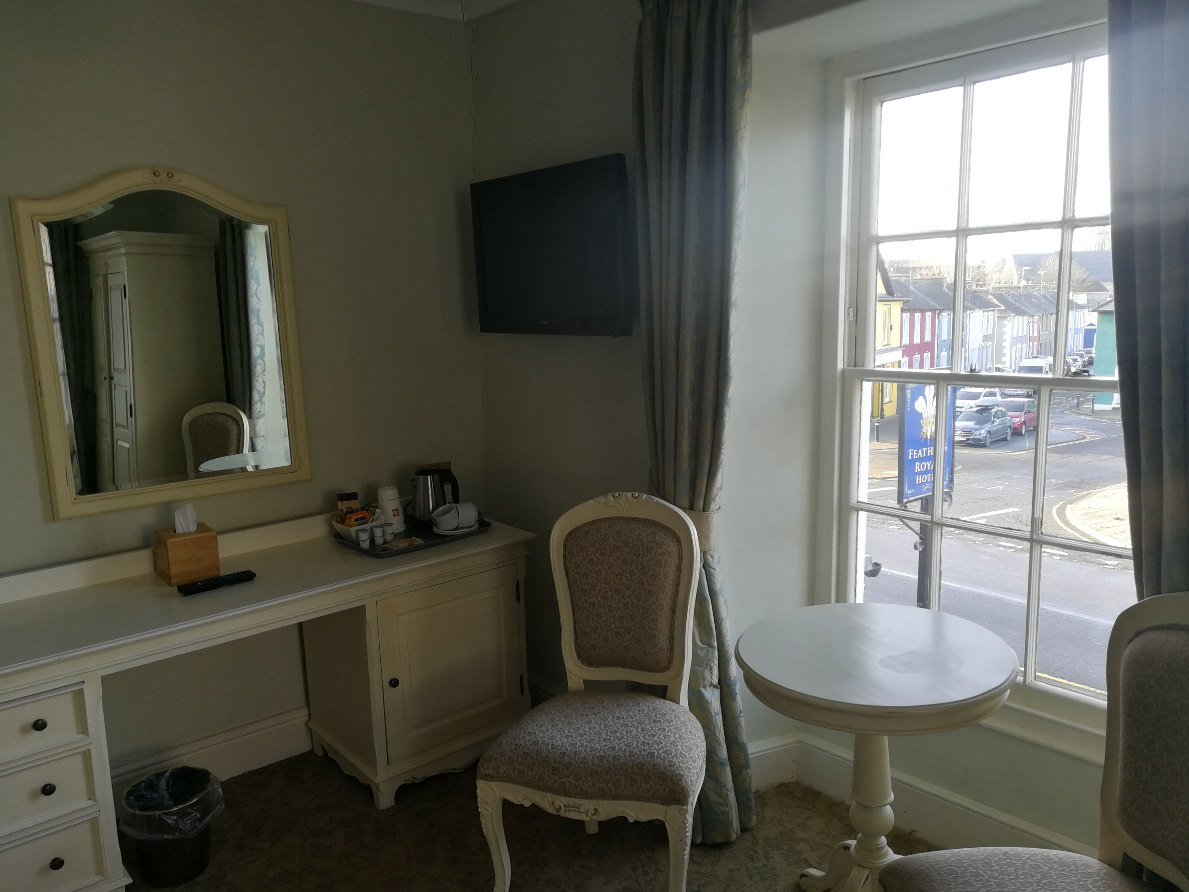 Een hotelkamer met een bureau, een stoel, een klein tafeltje, een televisie en een raam met uitzicht op de straat.