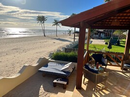 Un patio frente a la playa con vista al océano, palmeras y un espacio verde con mobiliario exterior.