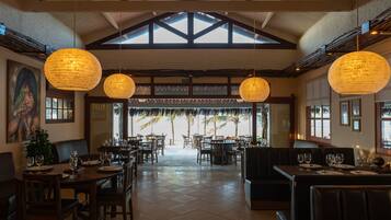 Restaurante com pé-direito alto, vigas aparentes e grandes luminárias pendentes. Mesas montadas com taças de vinho e pratos, com vista para palmeiras pela entrada.