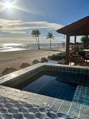 A pool area with a view of the beach, palm trees, and a thatched-roof structure.