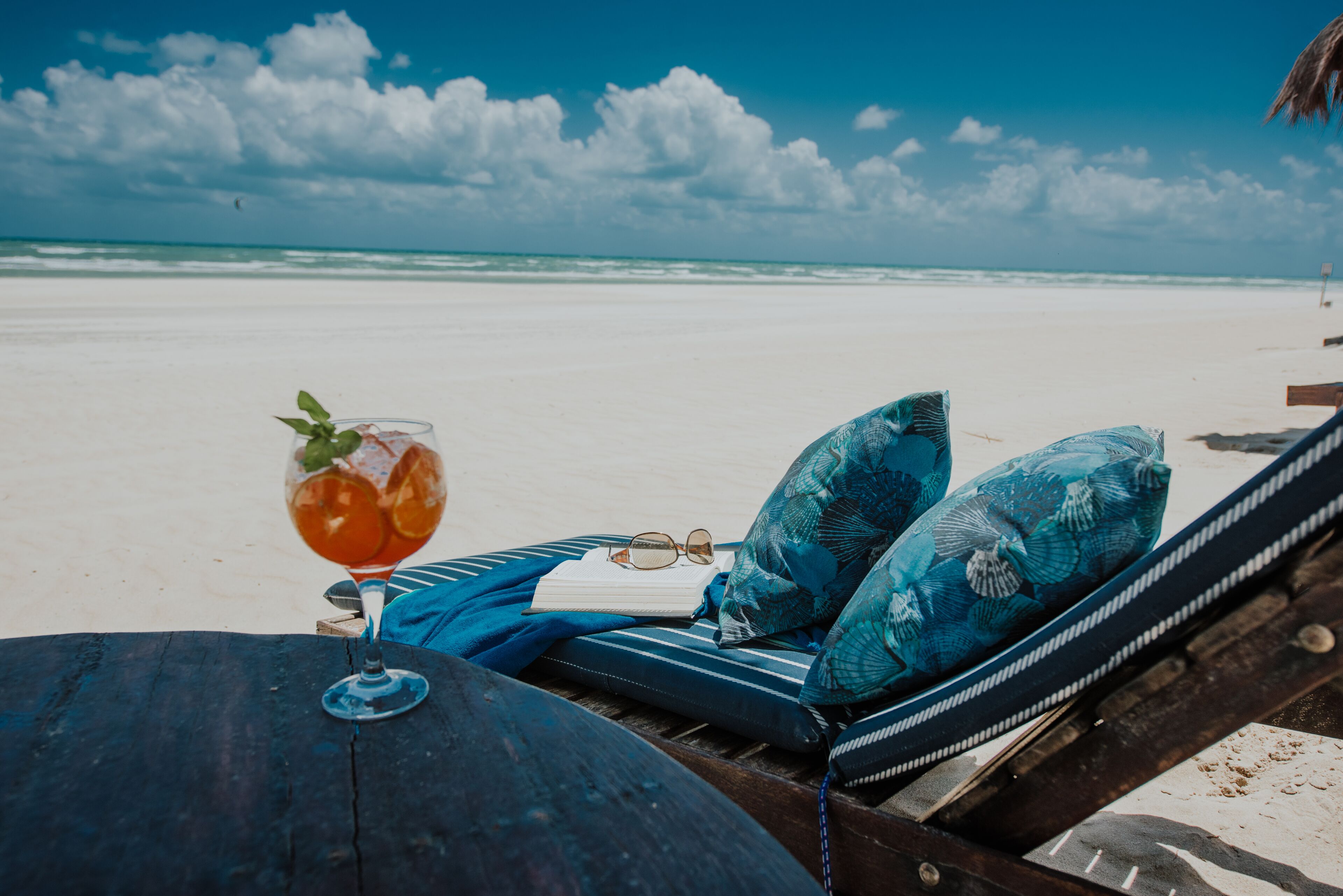 A beachside lounge chair with a glass of drink, a book, and a pair of sunglasses.