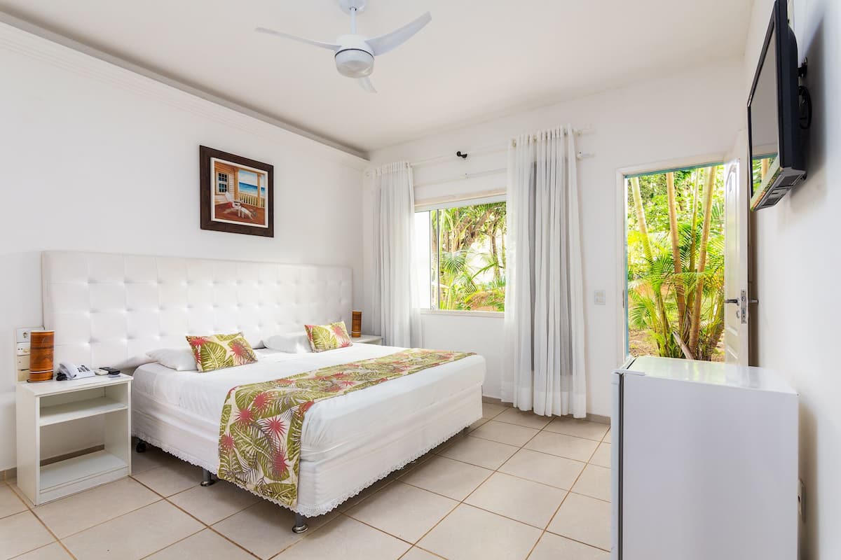 Une chambre d’hôtel avec un grand lit, une table de chevet, un ventilateur de plafond, une fenêtre avec des rideaux et un téléviseur fixé au mur.