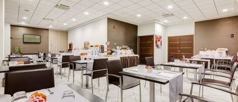 A modern dining area with multiple tables and chairs, a kitchen area in the background, and a flat-screen TV mounted on the wall.