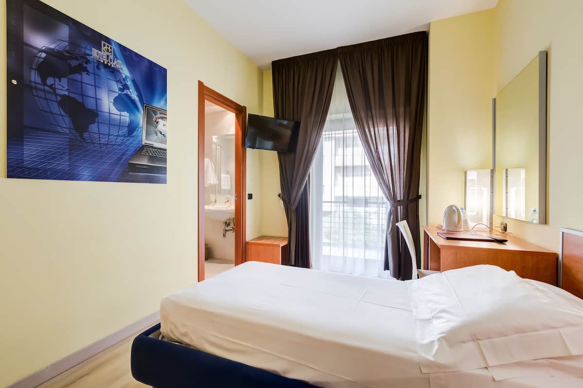 A hotel room with a bed, a desk, a chair, a TV, a window with curtains, and a bathroom visible through an open door.