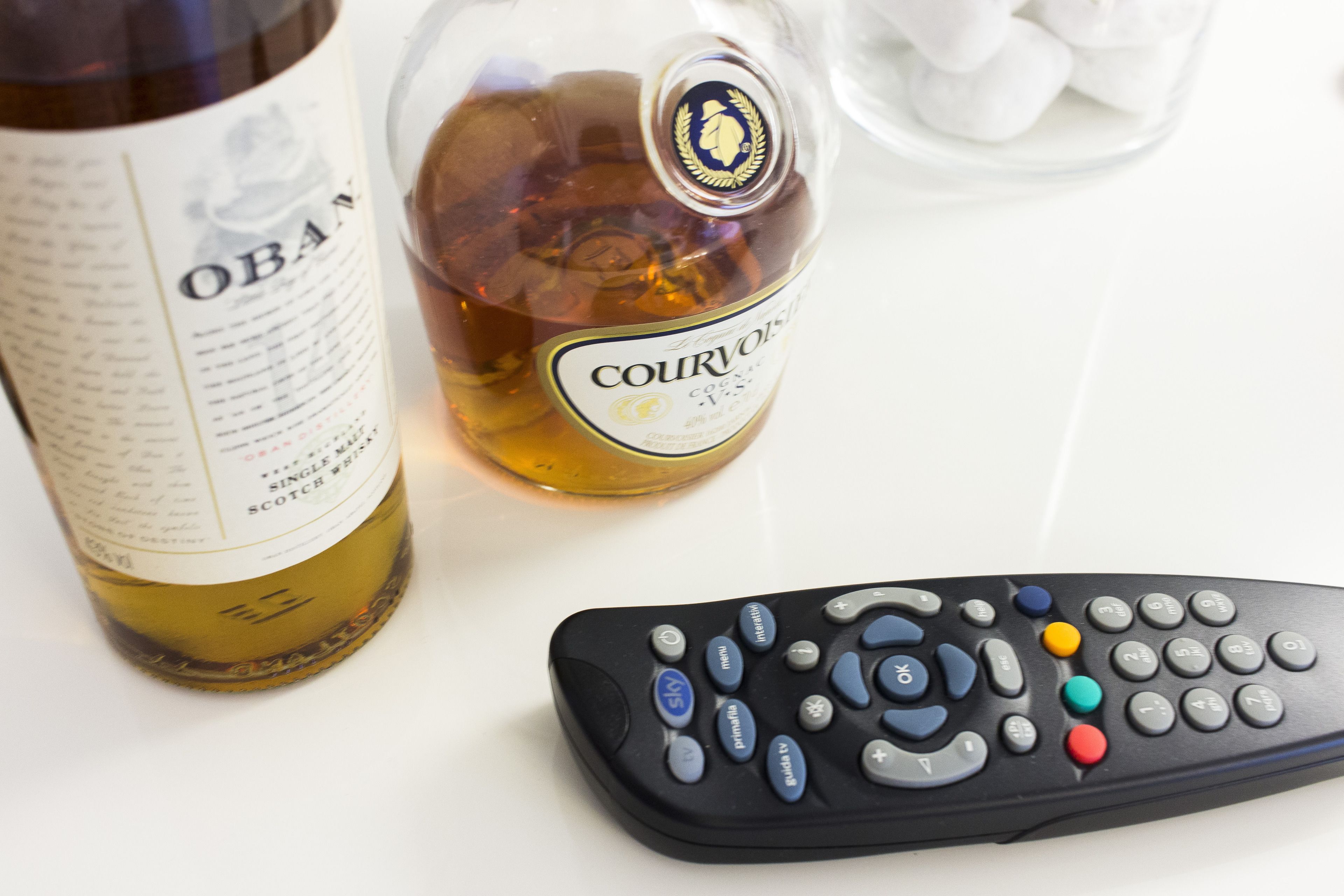 A remote control, a bottle of Courvoisier cognac, and a bottle of Oban whisky on a white surface.