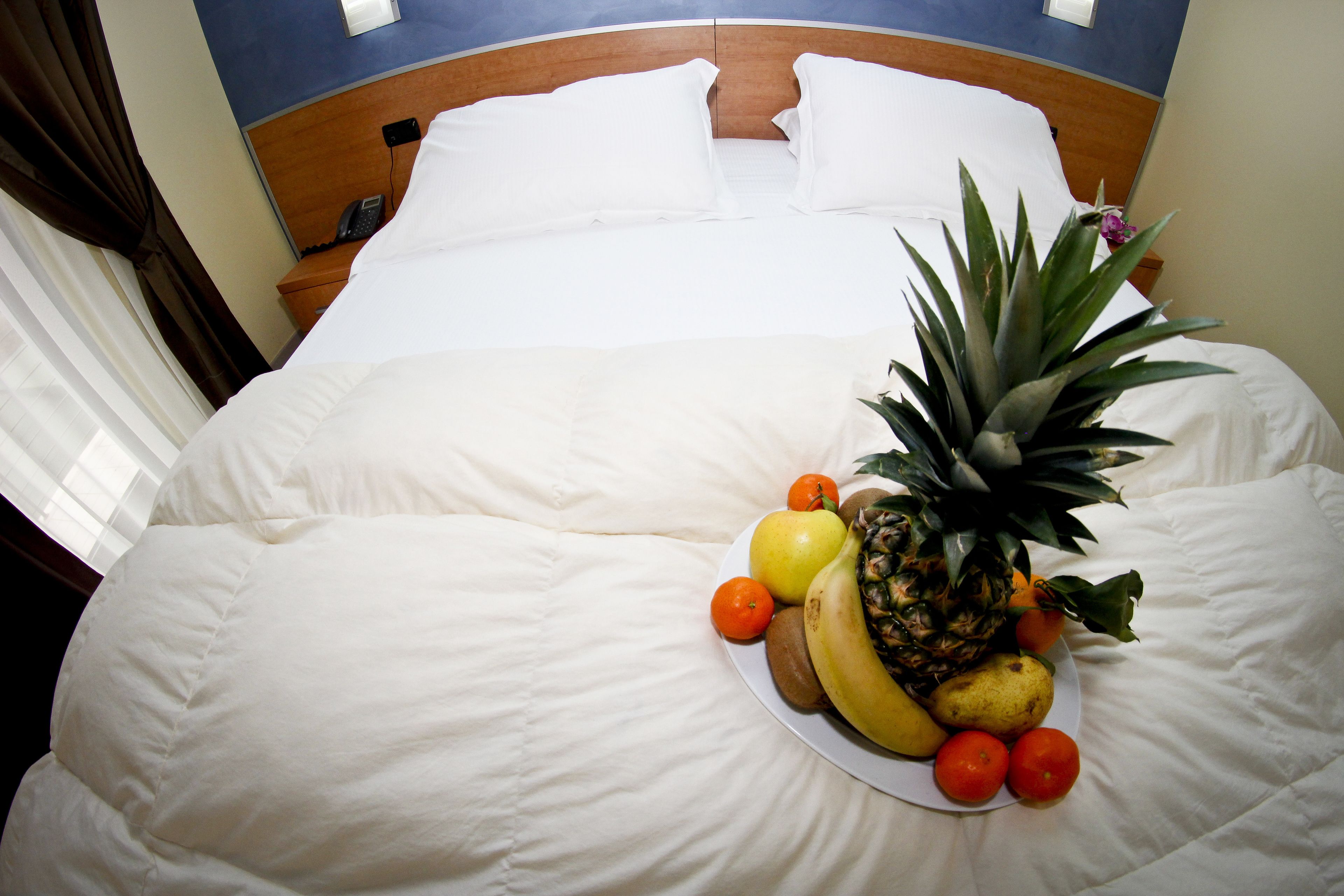 A neatly made bed with white linens, a fruit plate on the bed, and a wooden headboard.