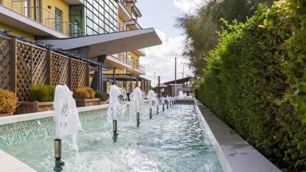Una piscina con múltiples chorros de agua, rodeada por una valla de madera y un seto.