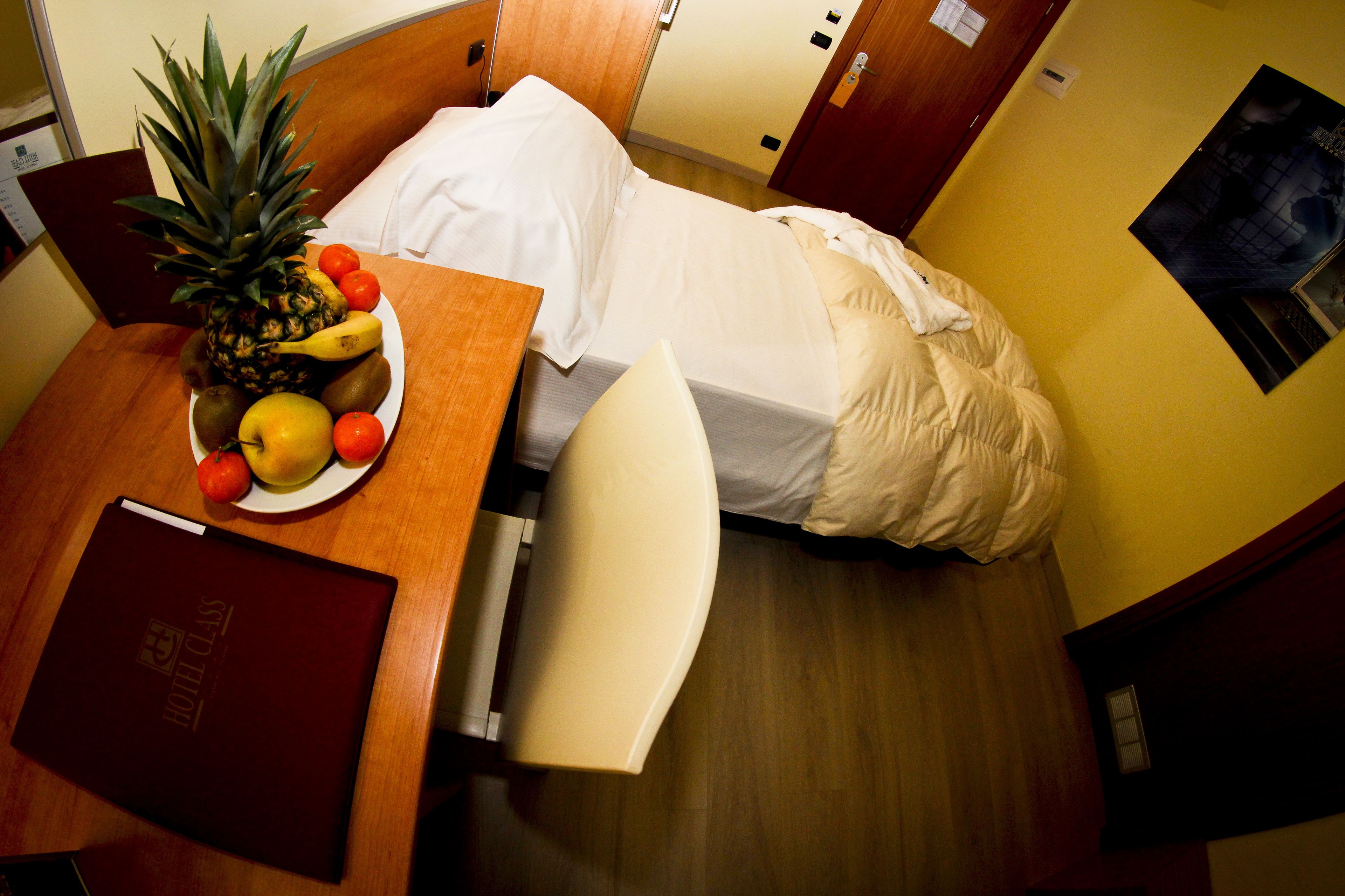 Una camera d'albergo con un letto, un comodino con una ciotola di frutta, una sedia e una scrivania in legno con un libro.