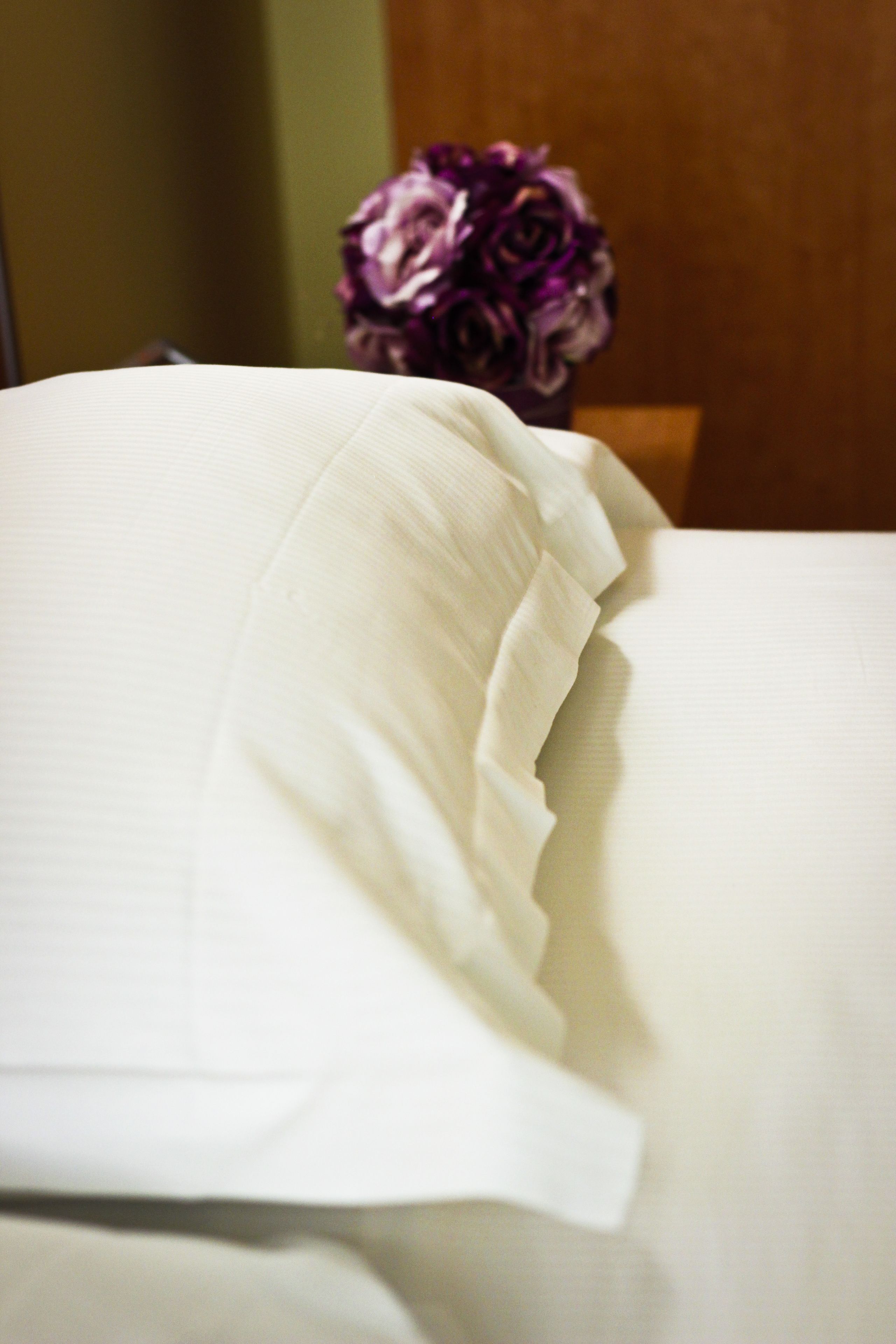 A bed with white linens and a bouquet of purple flowers in the background.