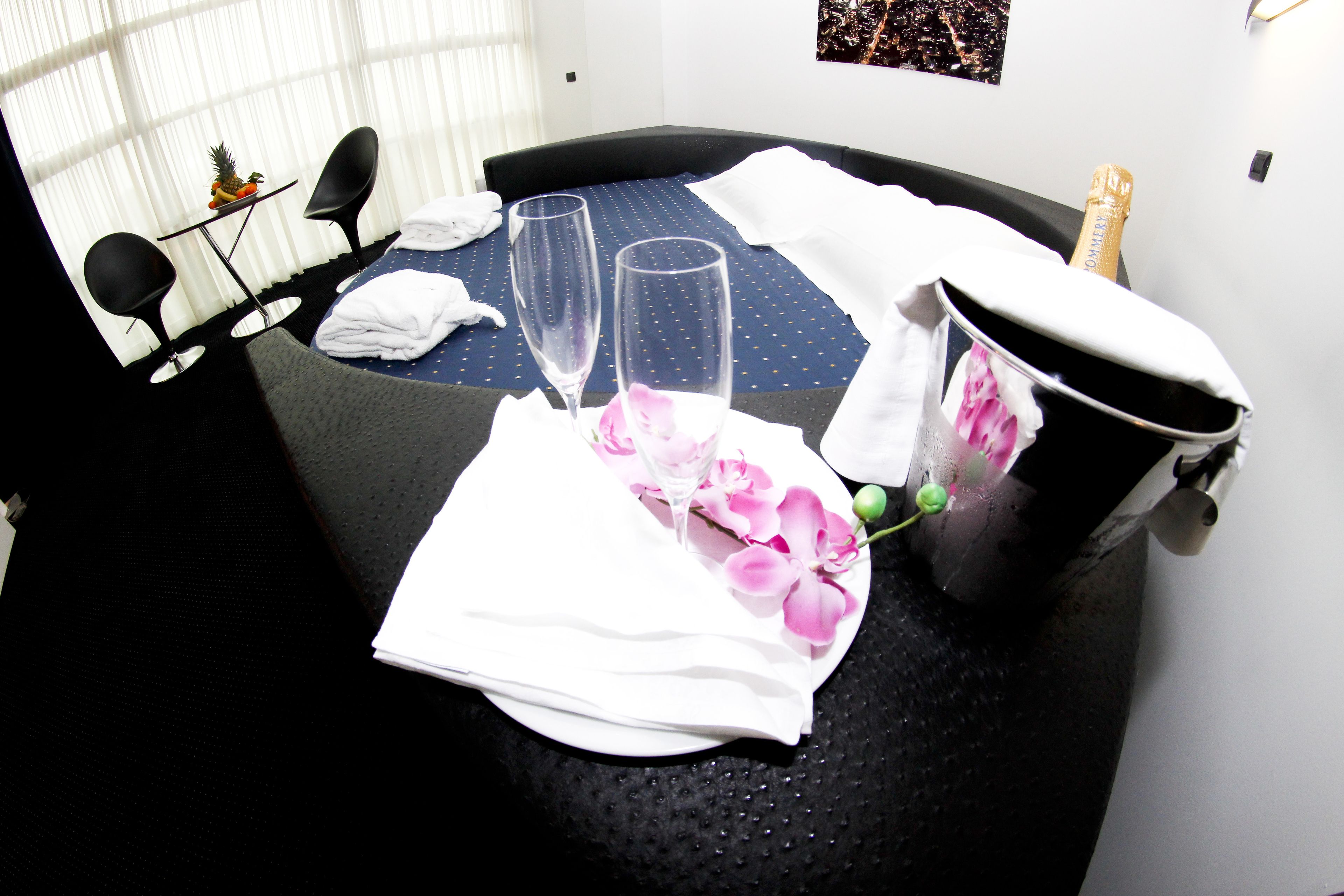 A neatly arranged hotel room with a bed, two chairs, a small table with a fruit bowl, two glasses, a bucket, and a bottle of champagne.