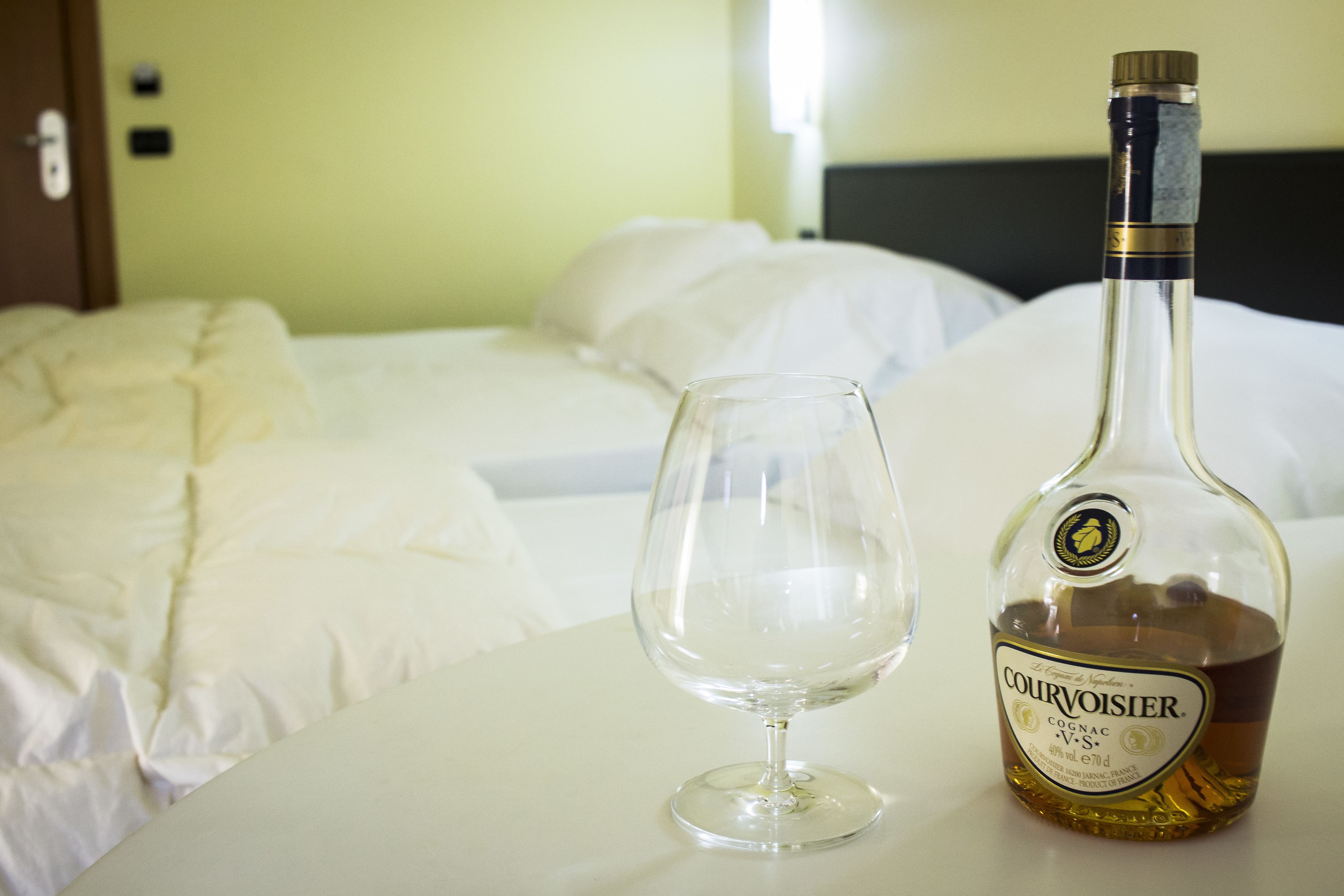 A bottle of Courvoisier cognac and an empty wine glass on a table in a hotel room with a bed and a nightstand.