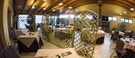 A spacious indoor dining area with multiple tables and chairs, wooden ceiling beams, and natural light from windows.