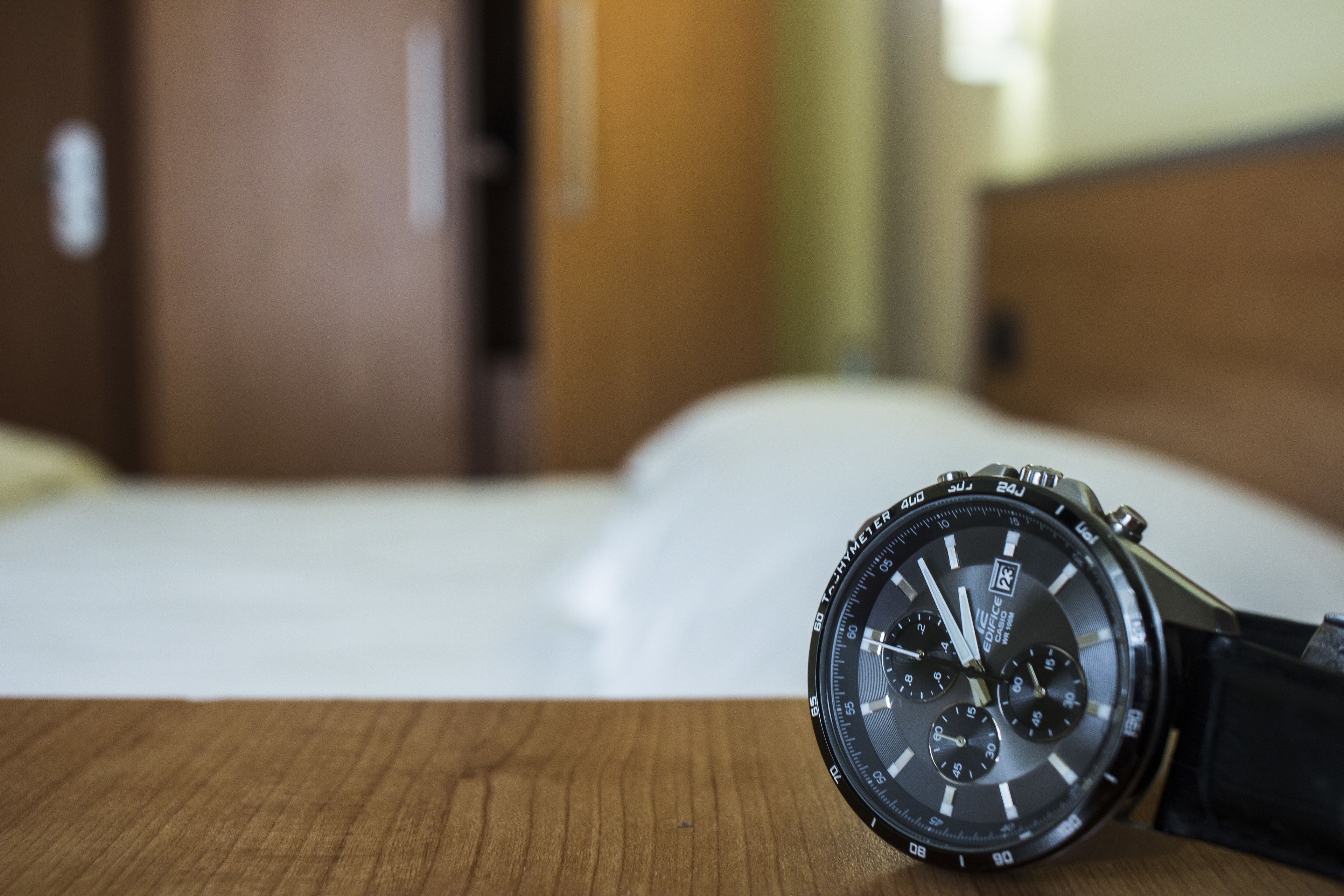 Eine Armbanduhr auf einer Holzoberfläche, im Hintergrund ein verschwommenes Bett und eine Tür.