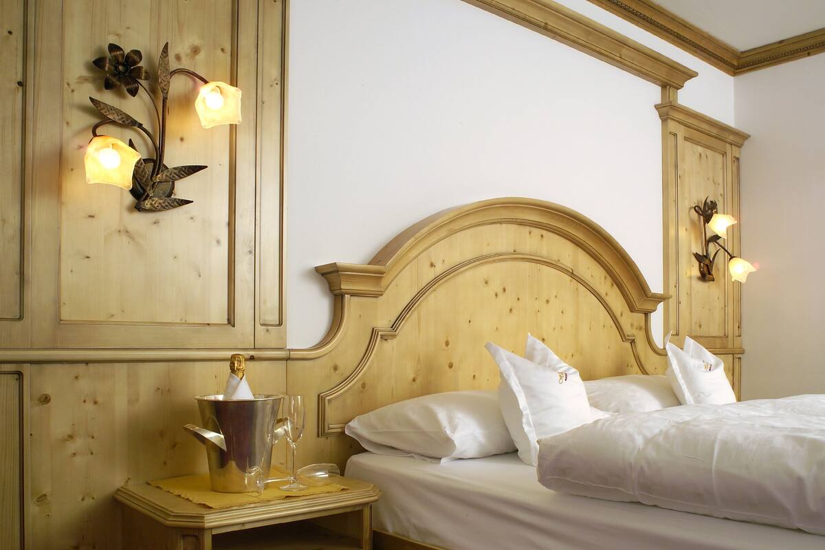 A wooden bed with white linens, a nightstand with a lamp, and wall-mounted sconces.