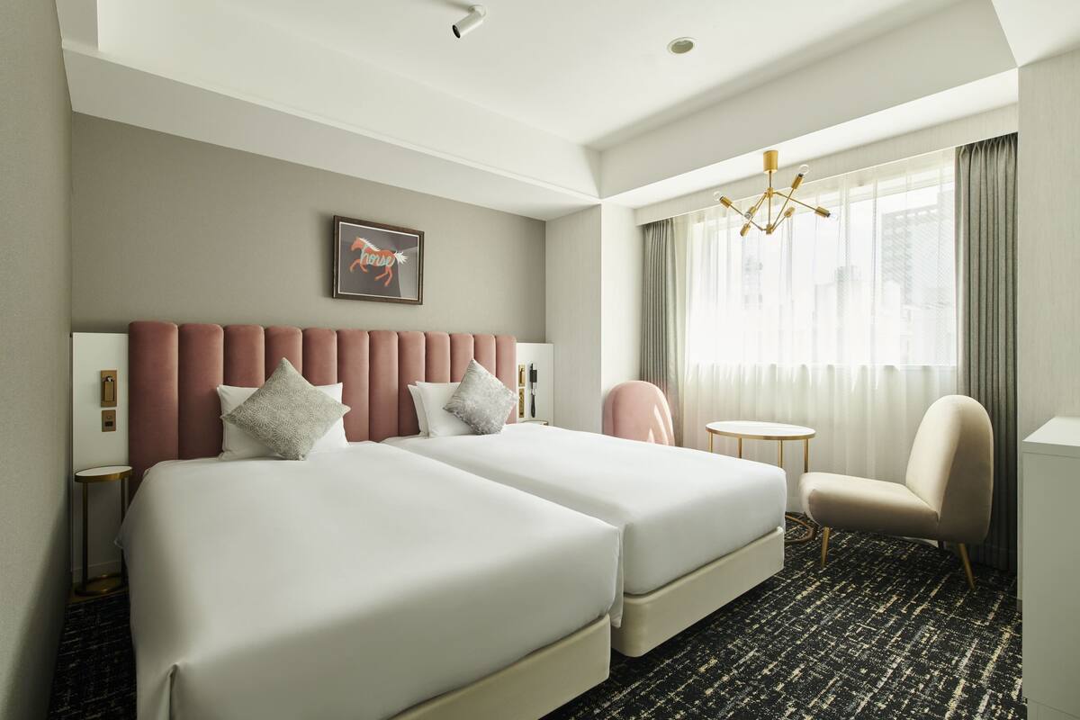 A hotel room with two beds, a chair, a small table, a chandelier, and a window with curtains.