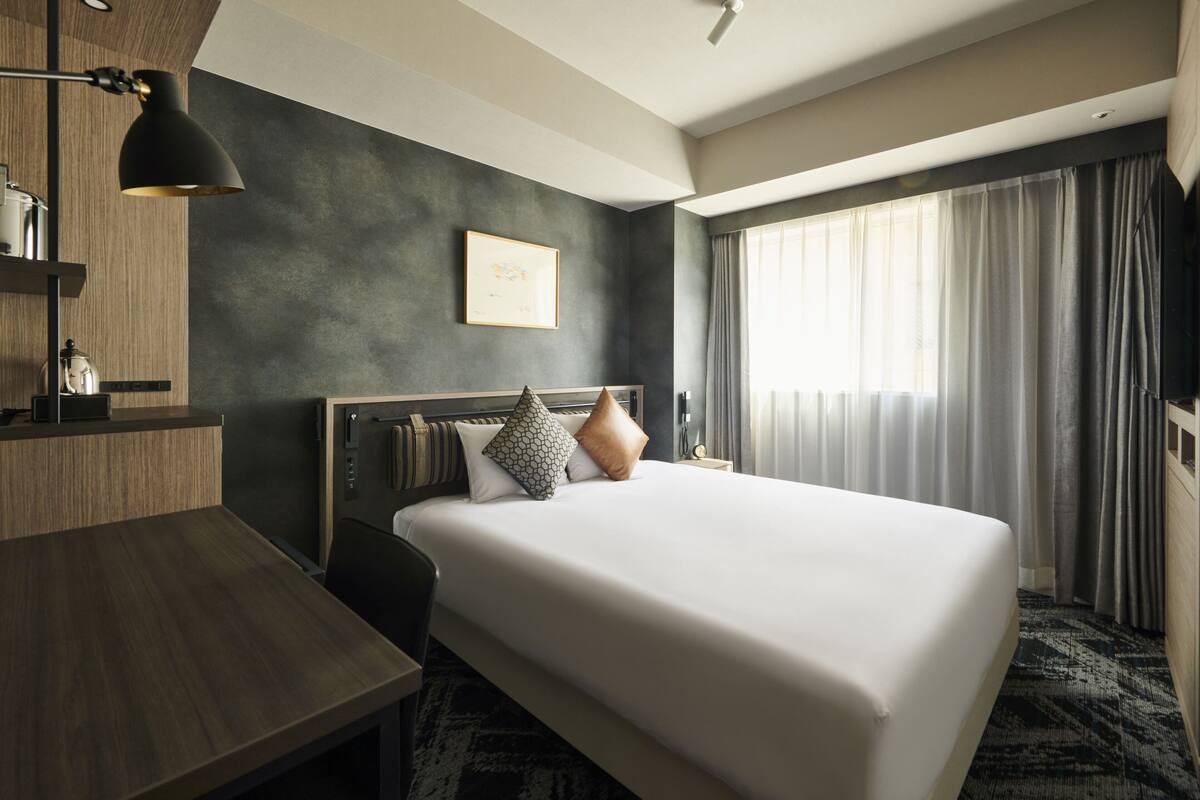 A hotel room with a large bed, a desk, a lamp, a framed picture on the wall, and a window with curtains.