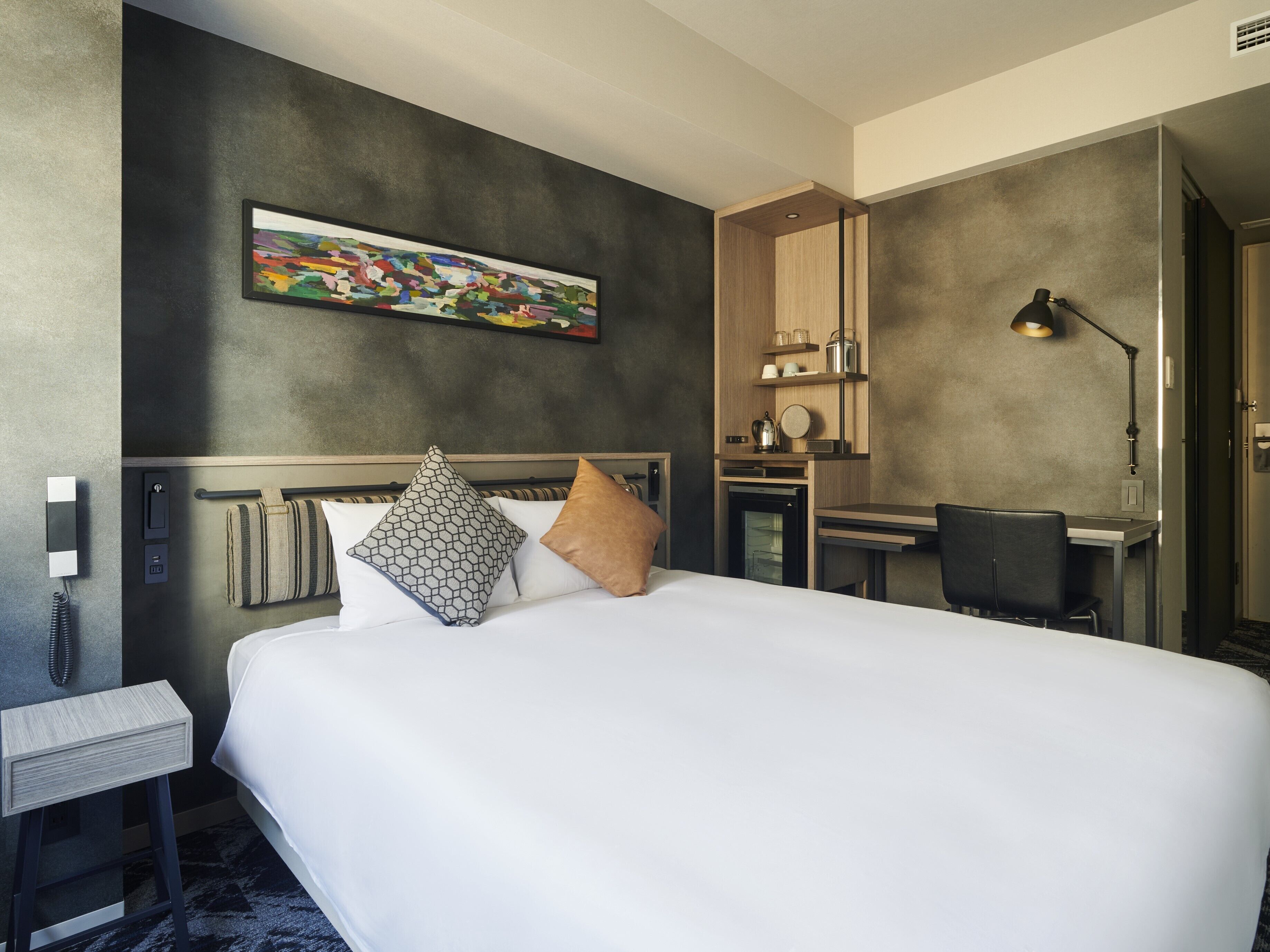 A hotel room with a large bed, a desk, a chair, a lamp, a painting on the wall, and a telephone.
