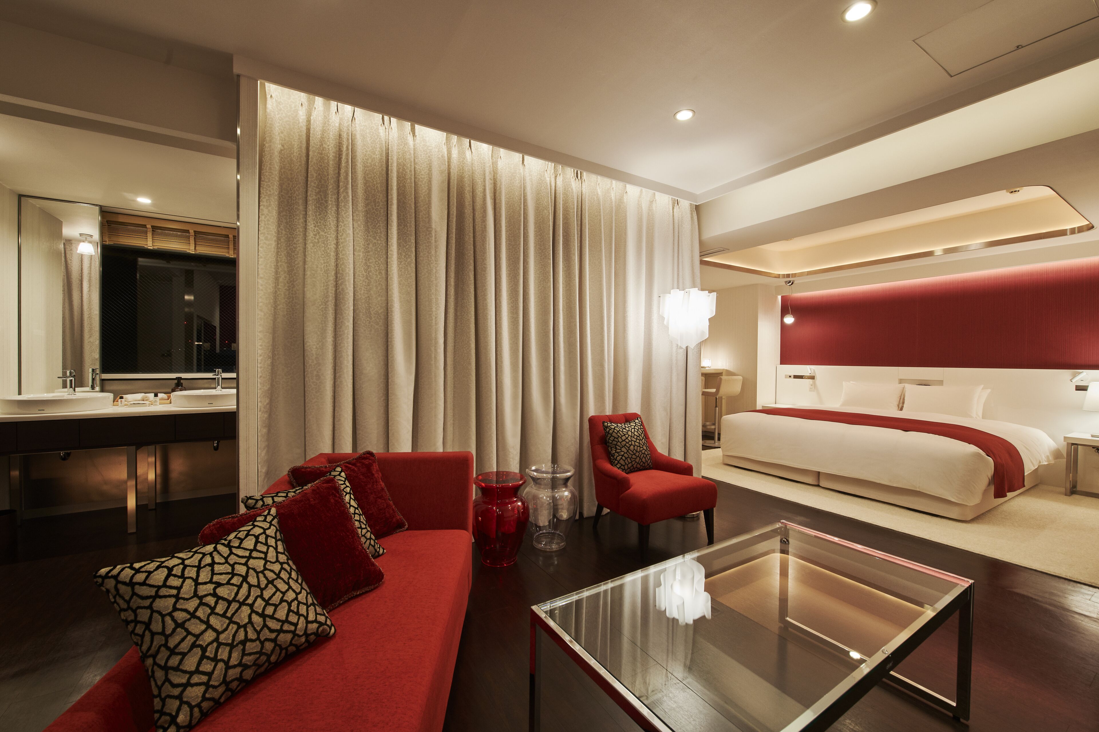 A modern hotel room with a red sofa, a glass coffee table, a bed with a red headboard, and a large mirror.