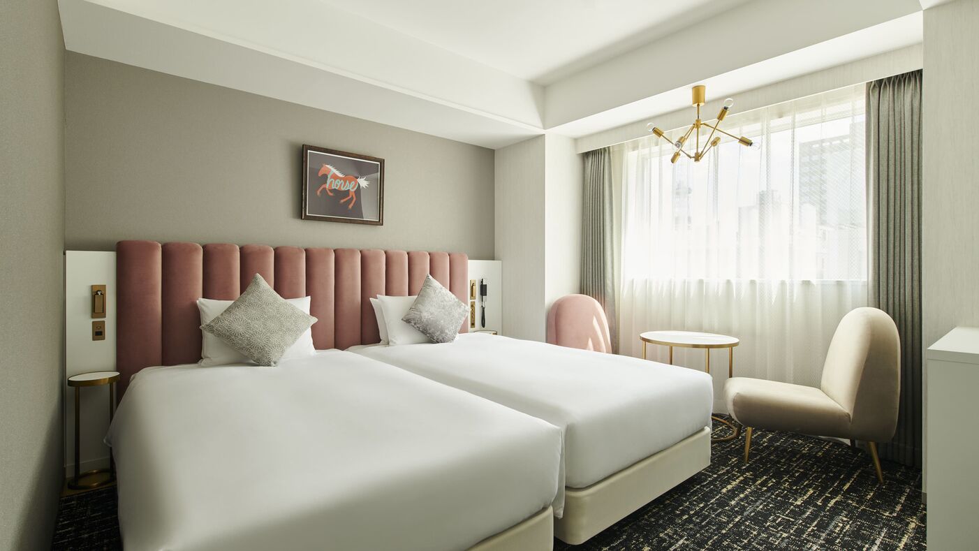 A hotel room with two beds, a chair, a small table, a chandelier, and a window with curtains.