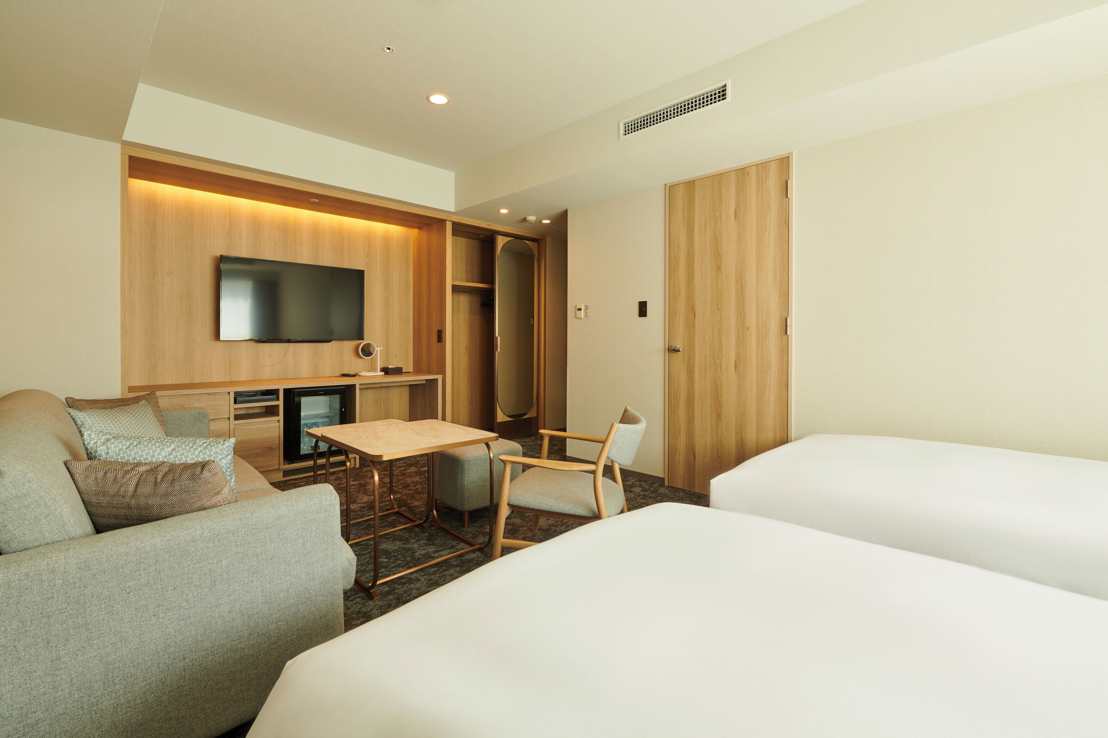 A hotel room with a flat-screen TV mounted on the wall, a wooden desk, a chair, a sofa, and two beds.