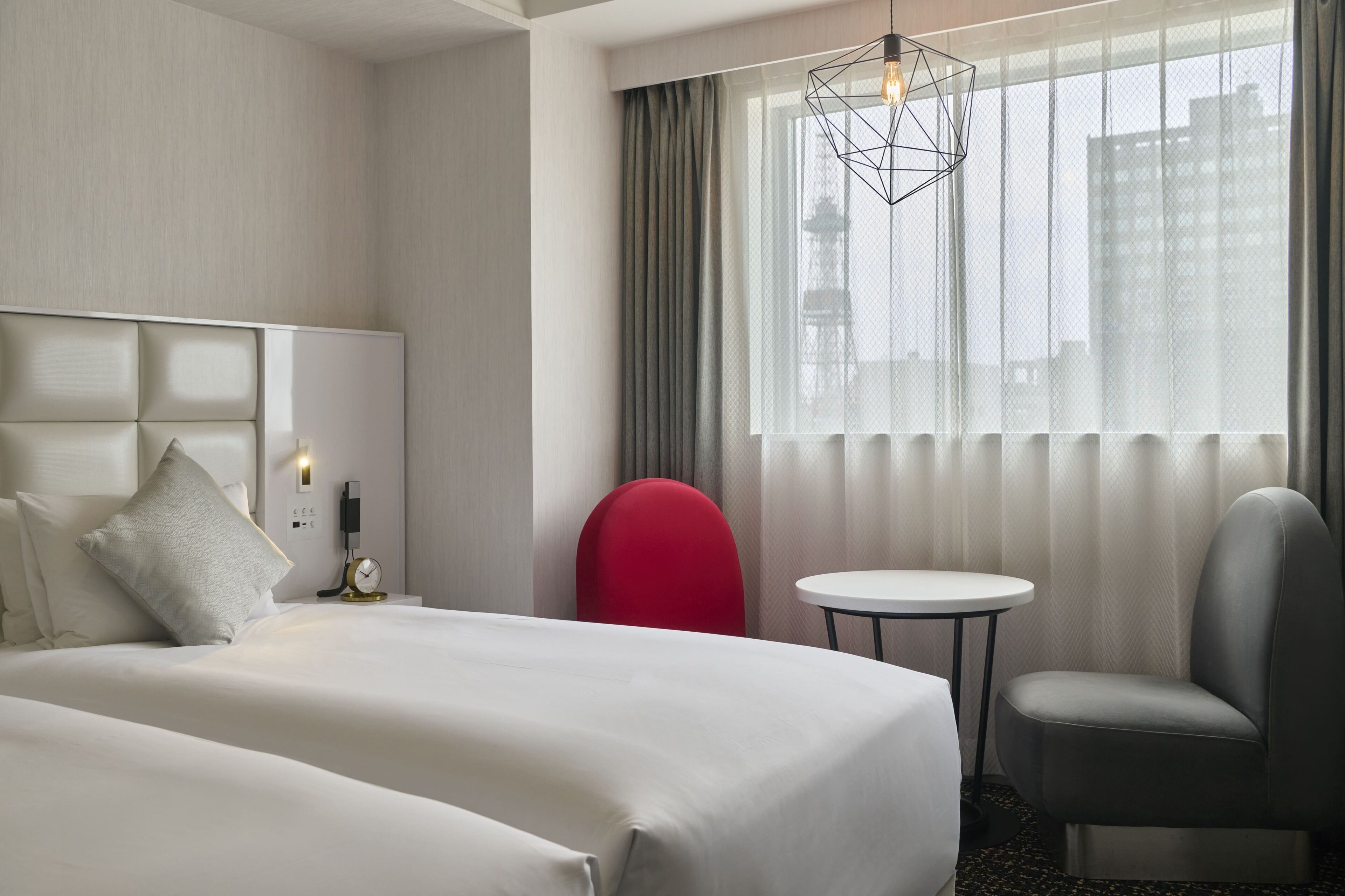A hotel room with a bed, a red chair, a small table, and a window with curtains.