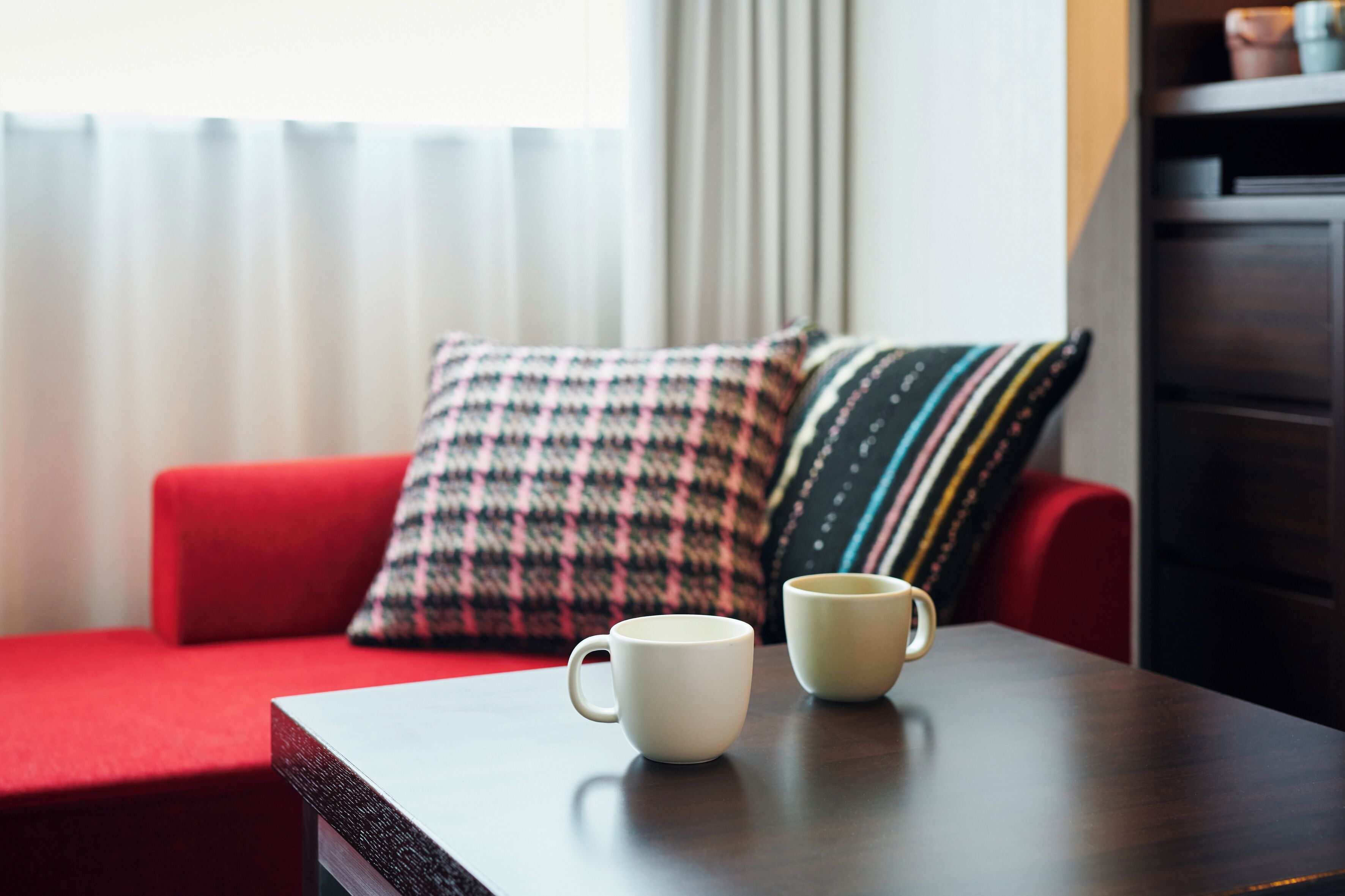 A room with a wooden table, two white mugs, a red sofa, and patterned cushions.