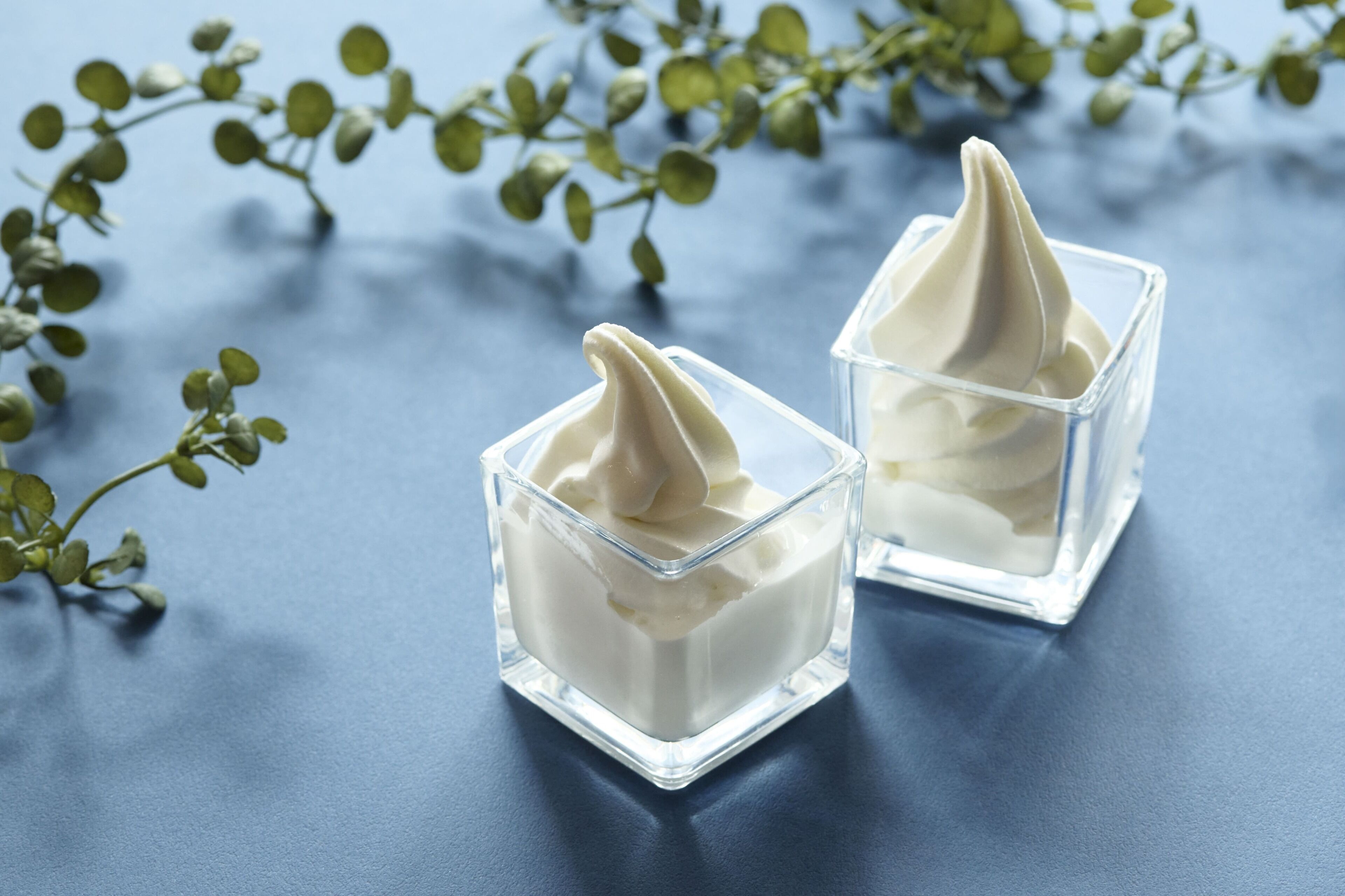 Dos recipientes de vidrio transparente con un contenido blanco, posiblemente un postre, sobre una superficie azul con follaje verde al fondo.